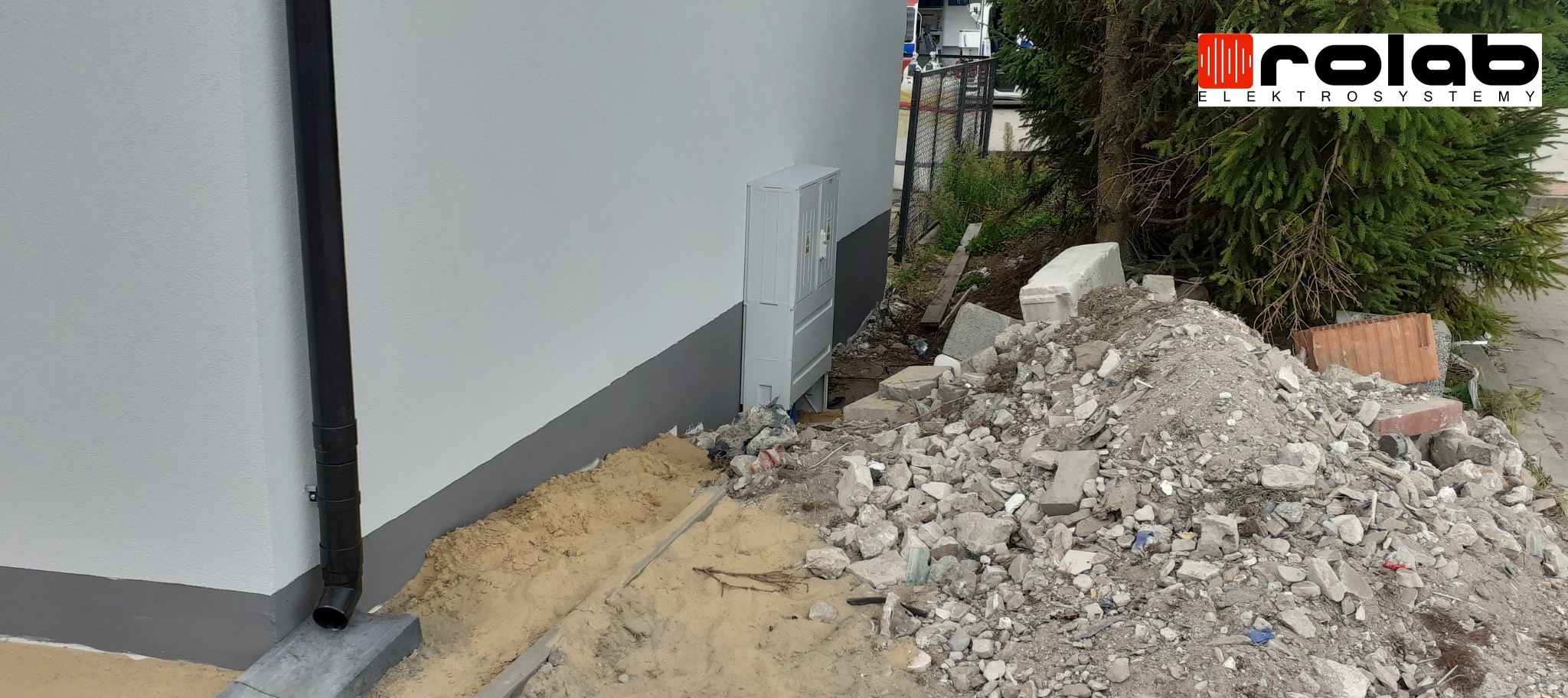 Szara skrzynka elektryczna na zewnątrz budynku, obok sterta gruzu i piasku, widoczny czarny spust rynnowy i logo firmy na tle drzewa.
