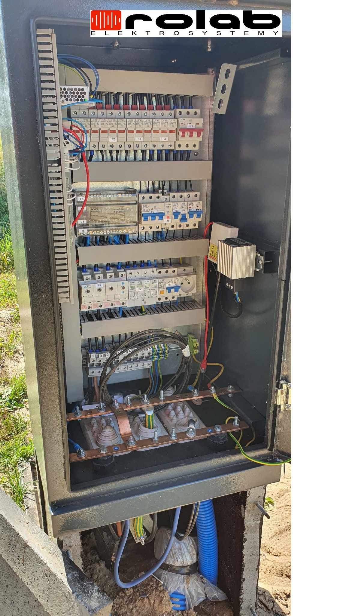 Otwarta szafa elektryczna zewnętrzna z widocznymi bezpiecznikami, transformatorami i okablowaniem, umieszczona w gruncie przy betonowym obrzeżu.