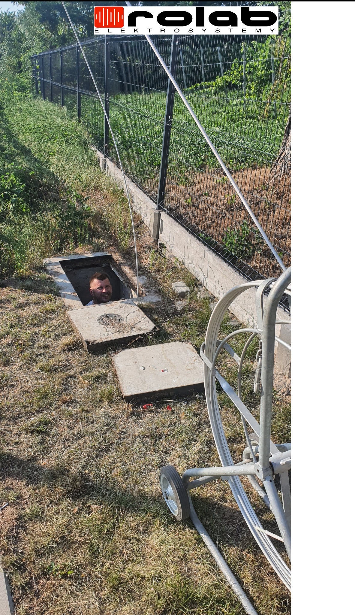 Elektryk wystający z otwartej studzienki kablowej podczas przeciągania kabla w pobliżu ogrodzenia z siatki, na trawie leżą betonowe pokrywy włazu, widoczny wózek z nawiniętym kablem.