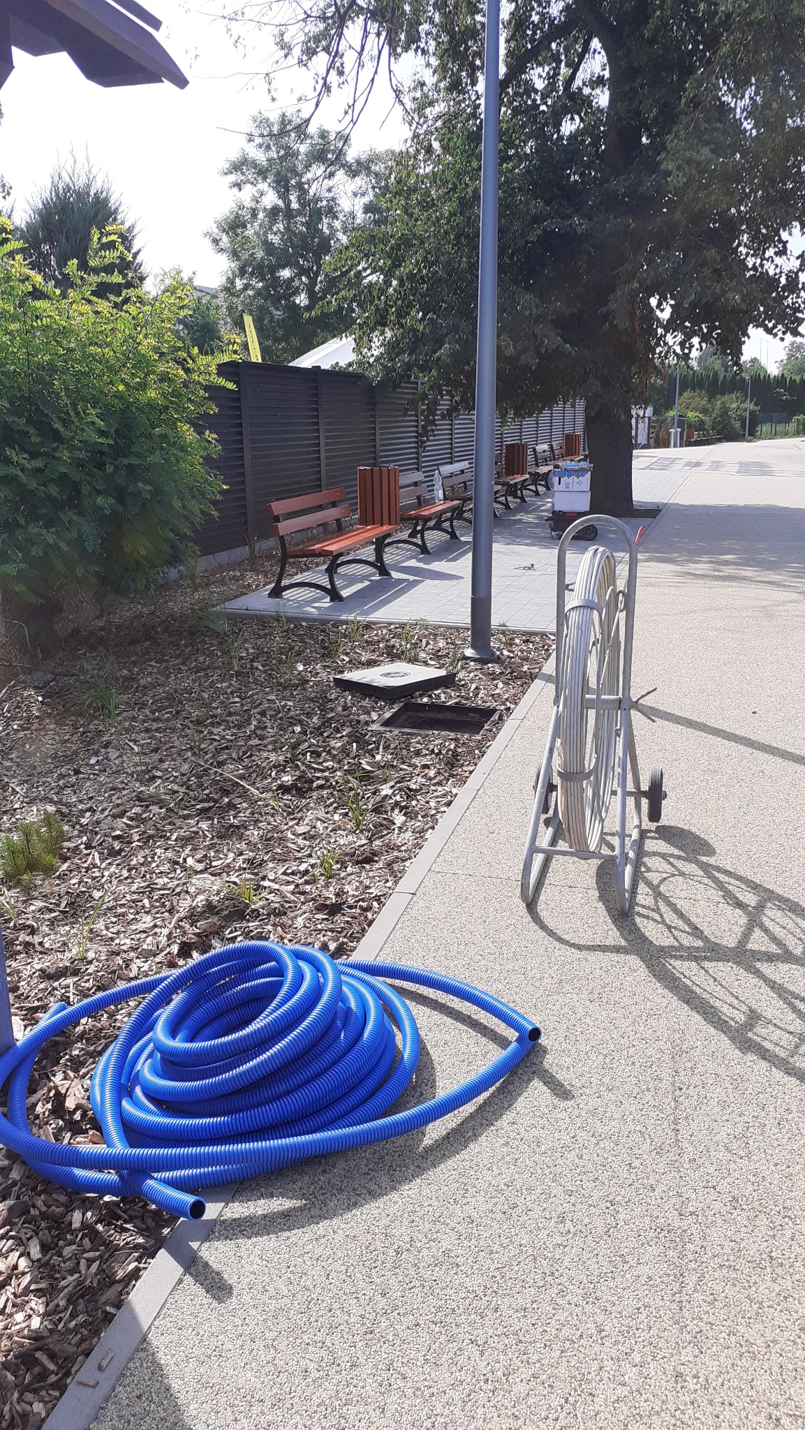 Niebieski karbowany przewód ułożony obok studzienki rewizyjnej na terenie zielonym w Iłży, obok zwinięty kabel na wózku.