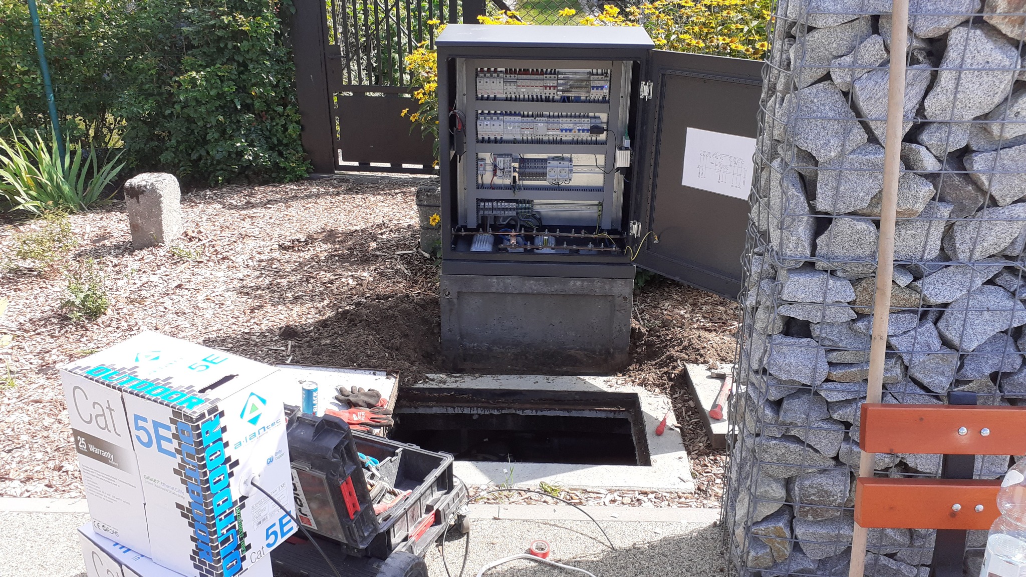 Otwarta szafa elektryczna z rozdzielnicą, skrzynka kablowa pod ziemią, narzędzia i kable Cat 5E na placu budowy, obok ogrodzenia gabionowego.