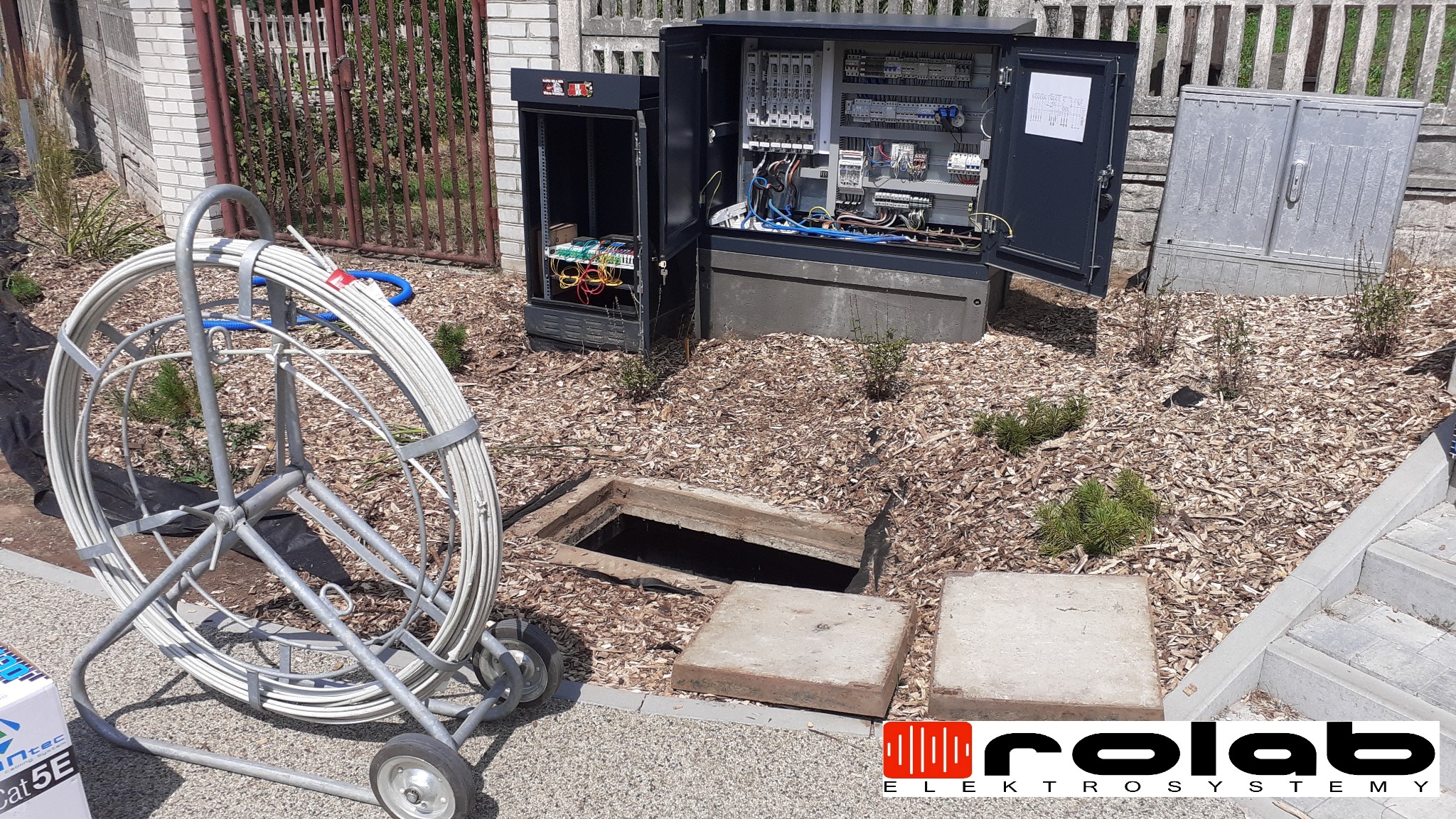 Otwarta szafa sterownicza z widocznymi przewodami i elementami elektrycznymi, bęben z kablem na wózku obok studzienki kanalizacyjnej z otwartym włazem, teren wokół wyłożony korą.