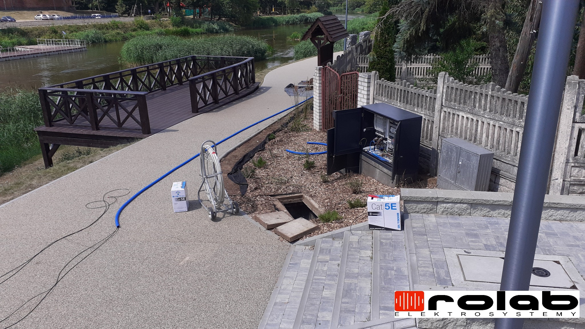 Otwarta szafa elektryczna z okablowaniem, skrzynki z kablem Cat5e, rura ochronna i studzienka kanalizacyjna na chodniku wzdłuż rzeki.
