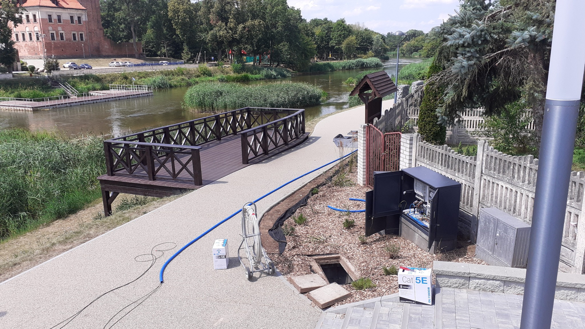 Otwarta szafka telekomunikacyjna z okablowaniem na tle ścieżki spacerowej nad rzeką, widoczny przewód telekomunikacyjny, skrzynka Cat5e i właz kanalizacyjny.