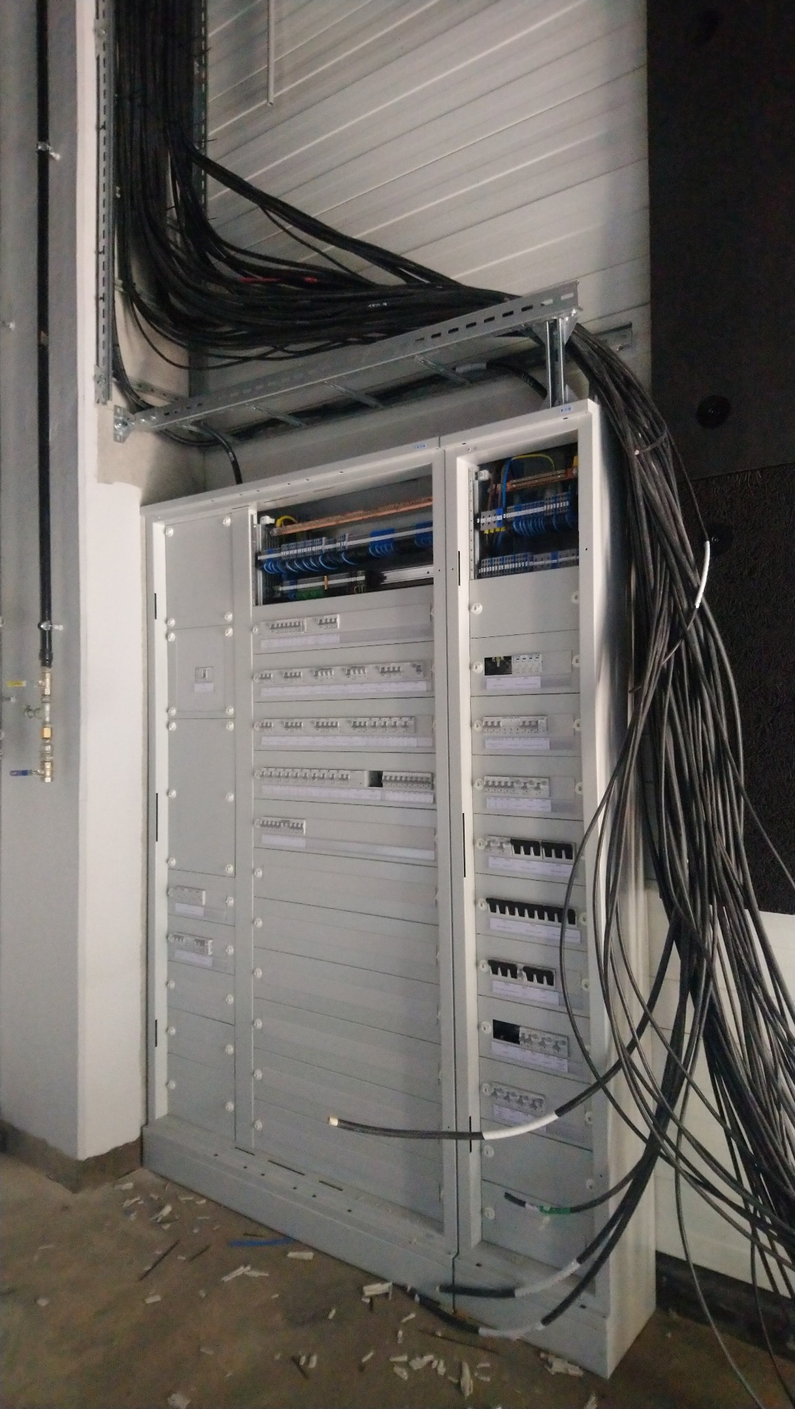 Rozbudowana szafa elektryczna z licznymi zabezpieczeniami i przewodami, widoczne kable prowadzone w korytach nad szafą, podłoga z drobnymi śmieciami budowlanymi.