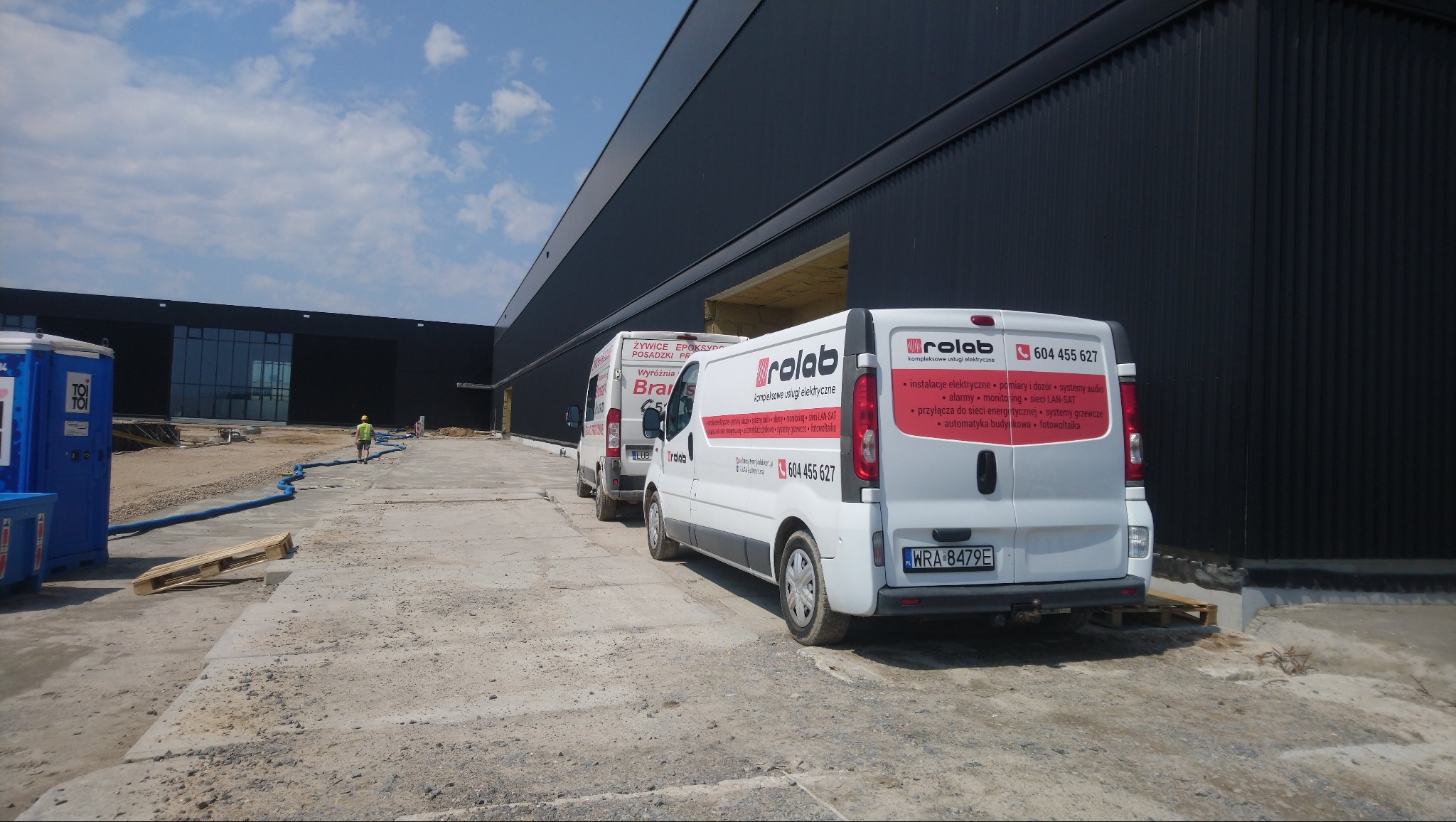 Dwa białe busy firmy Rolab z reklamą usług elektrycznych zaparkowane przed nowoczesnym budynkiem przemysłowym w trakcie dnia, widoczne przenośne toalety i pracownik w oddali.
