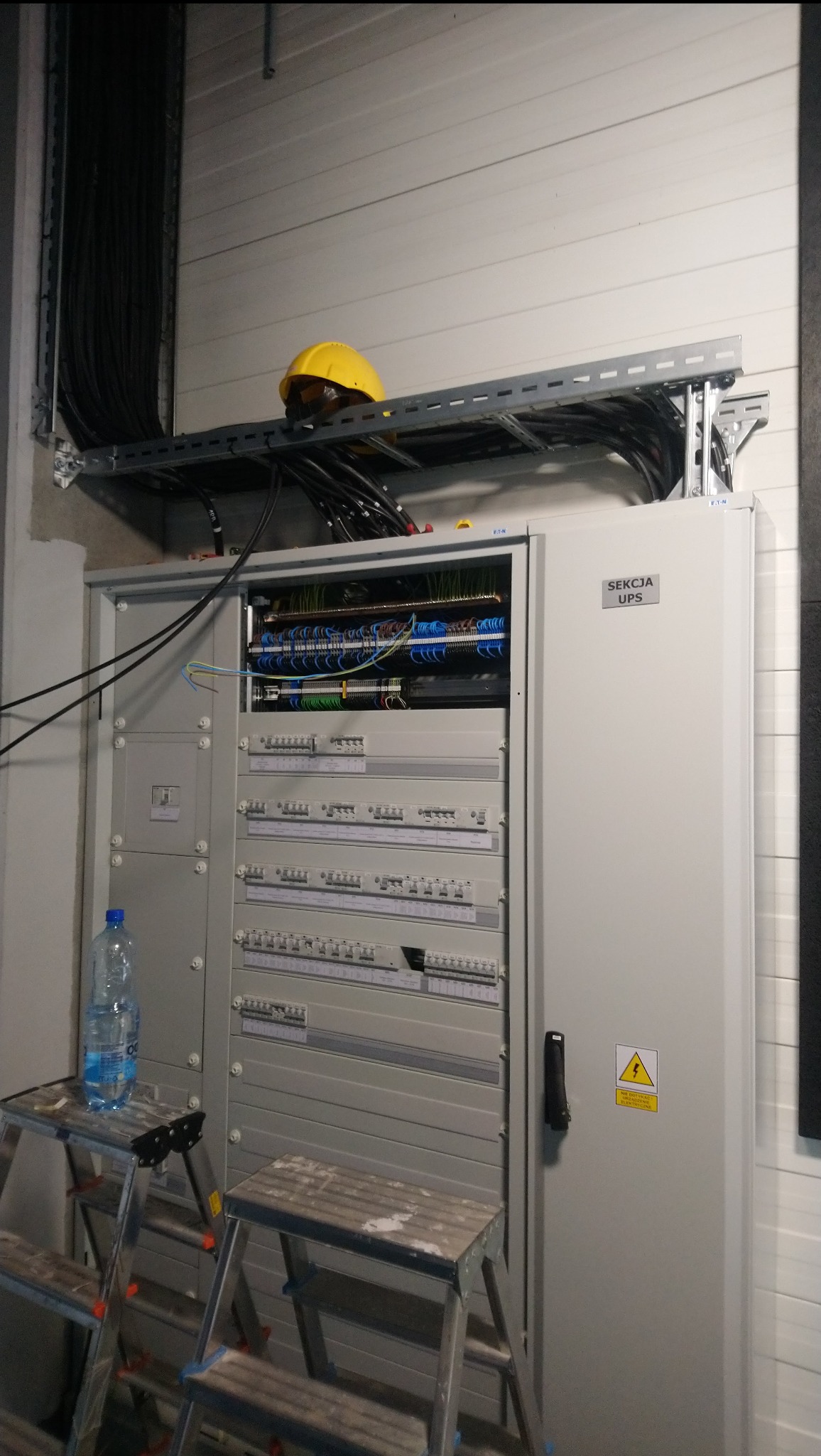 Szara szafa elektryczna UPS z otwartymi drzwiami, widoczne liczne przewody i złącza, żółty kask na półce powyżej, butelka wody na aluminiowej drabinie.