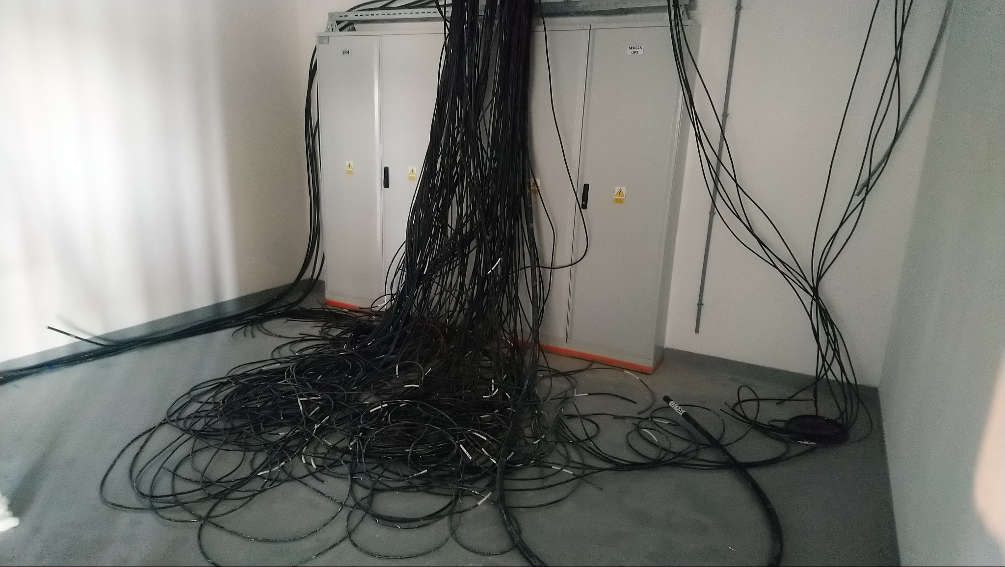 Stojące szafy elektryczne z nieuporządkowaną wiązką czarnych kabli na podłodze, widoczne oznaczenia na szafach i ostrzeżenie o napięciu.