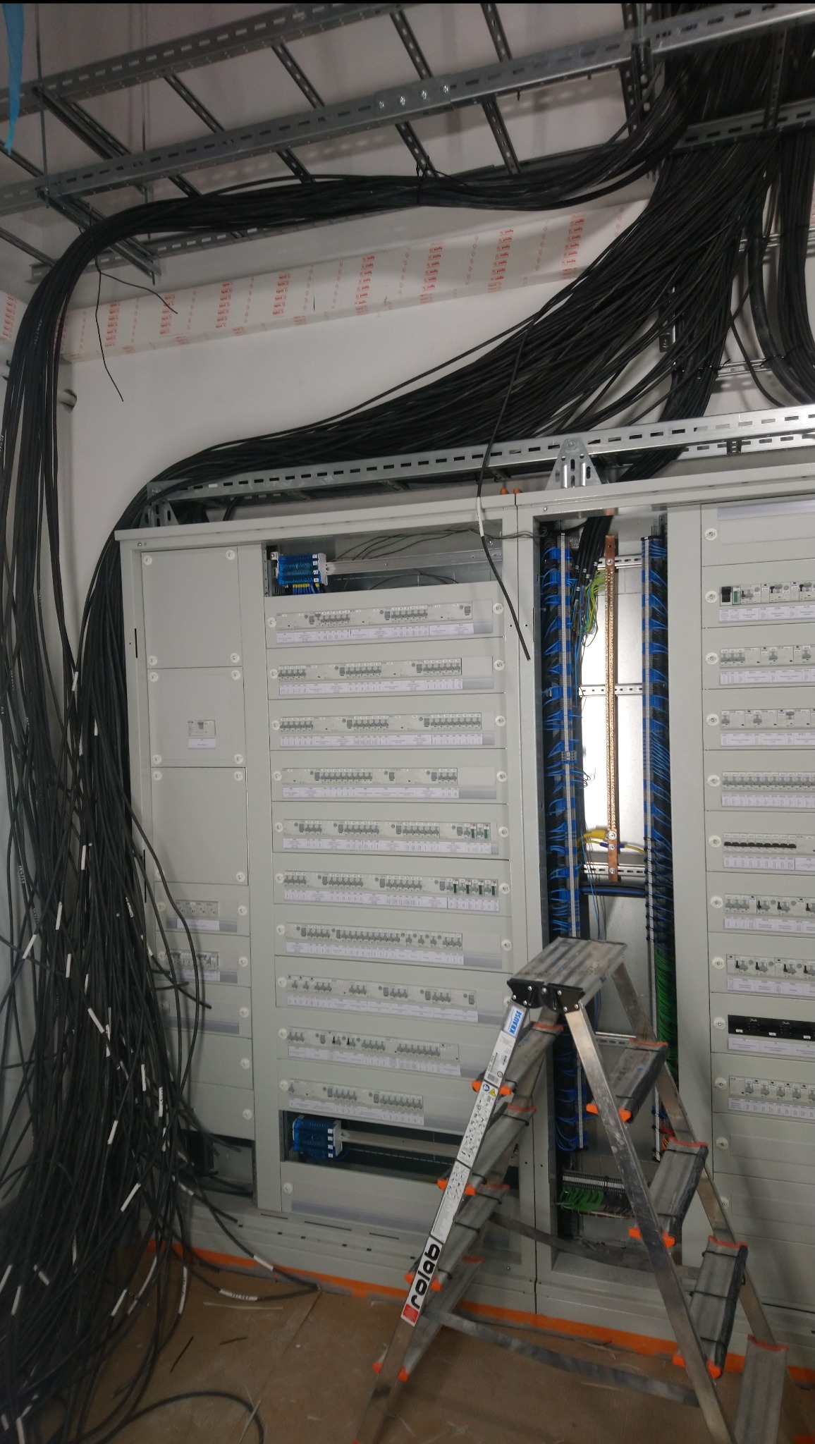 Rozdzielnica elektryczna w trakcie montażu, widoczne liczne kable, drabina aluminiowa oraz szyny montażowe pod sufitem.