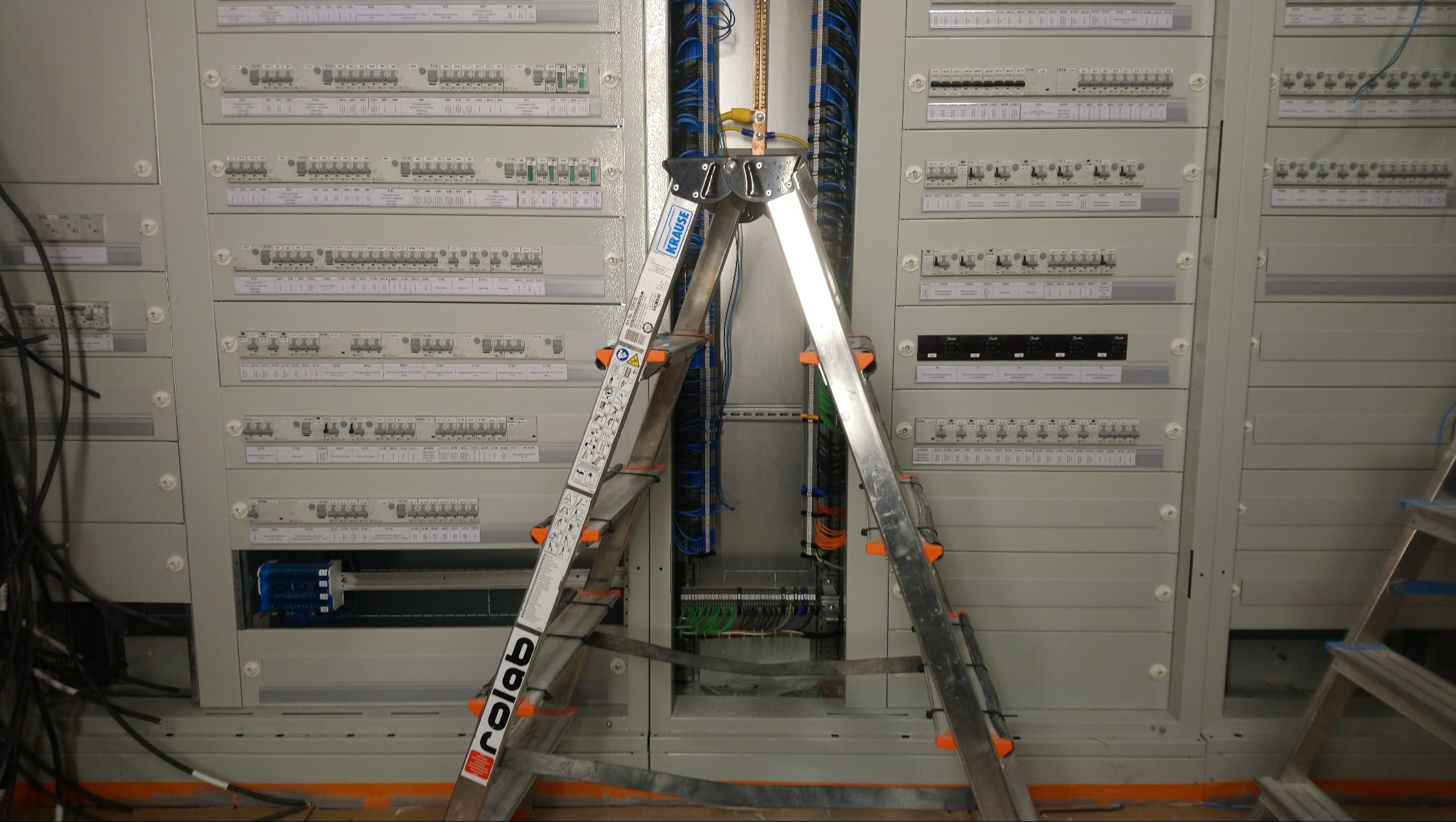 Rozdzielnica elektryczna z licznymi bezpiecznikami i okablowaniem, widoczna drabina aluminiowa z napisem Rolab.