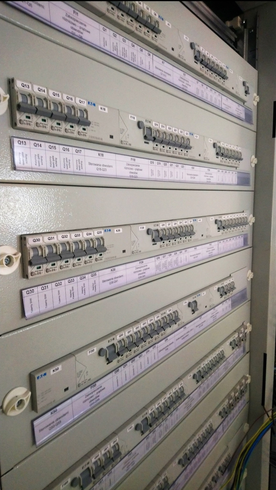 Rozdzielnica elektryczna z modułowymi wyłącznikami i opisami obwodów, widoczne etykiety z oznaczeniami Q1-Q6, Q13-Q18, Q30-Q35, K50, F150,  w tle widoczne kable.