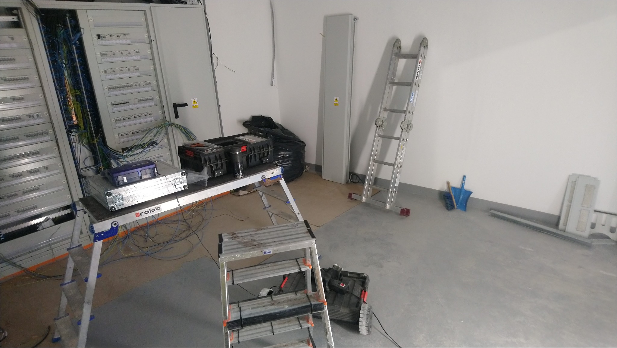 Rozdzielnica elektryczna z podłączonymi przewodami, narzędzia i drabiny w pomieszczeniu w trakcie prac instalacyjnych.