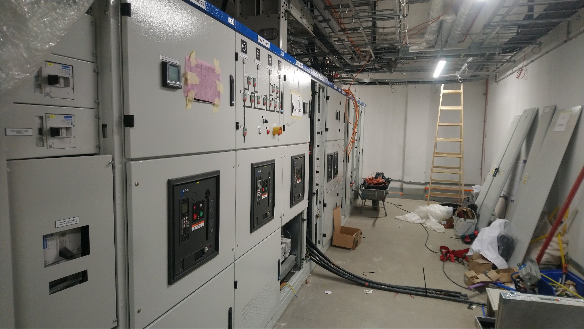 Rozbudowana rozdzielnica elektryczna w surowym pomieszczeniu z widocznymi kablami, osprzętem i drabiną, sugerująca etap instalacji.