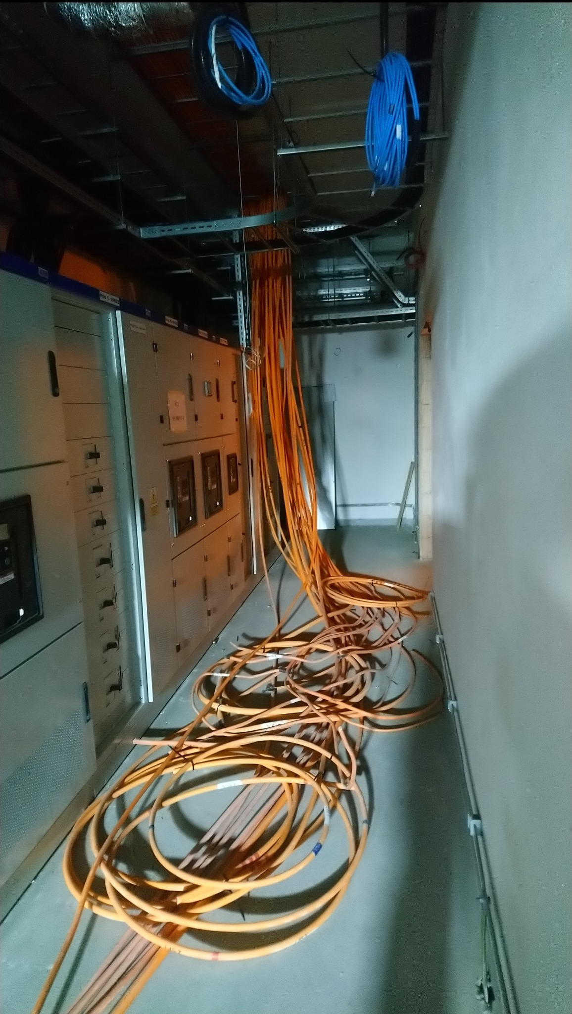 Rozłożone na podłodze pomarańczowe kable elektryczne obok szarej szafy sterowniczej z przełącznikami i miernikami, z niebieskimi kablami zwisającymi z sufitu