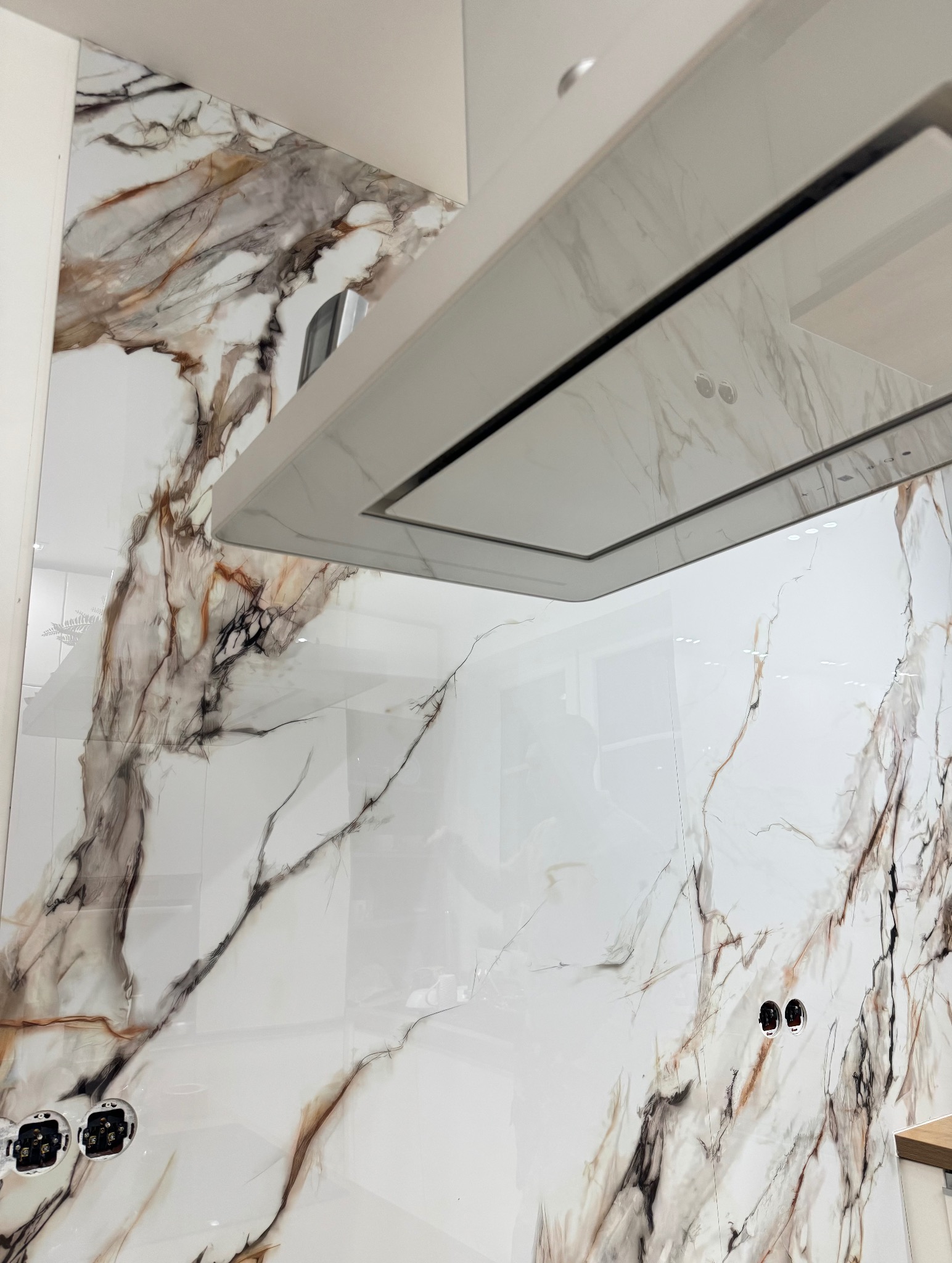 Białe płytki imitujące marmur z brązowymi żyłami na ścianie nad blatem kuchennym, widoczne gniazdka elektryczne i okap kuchenny.