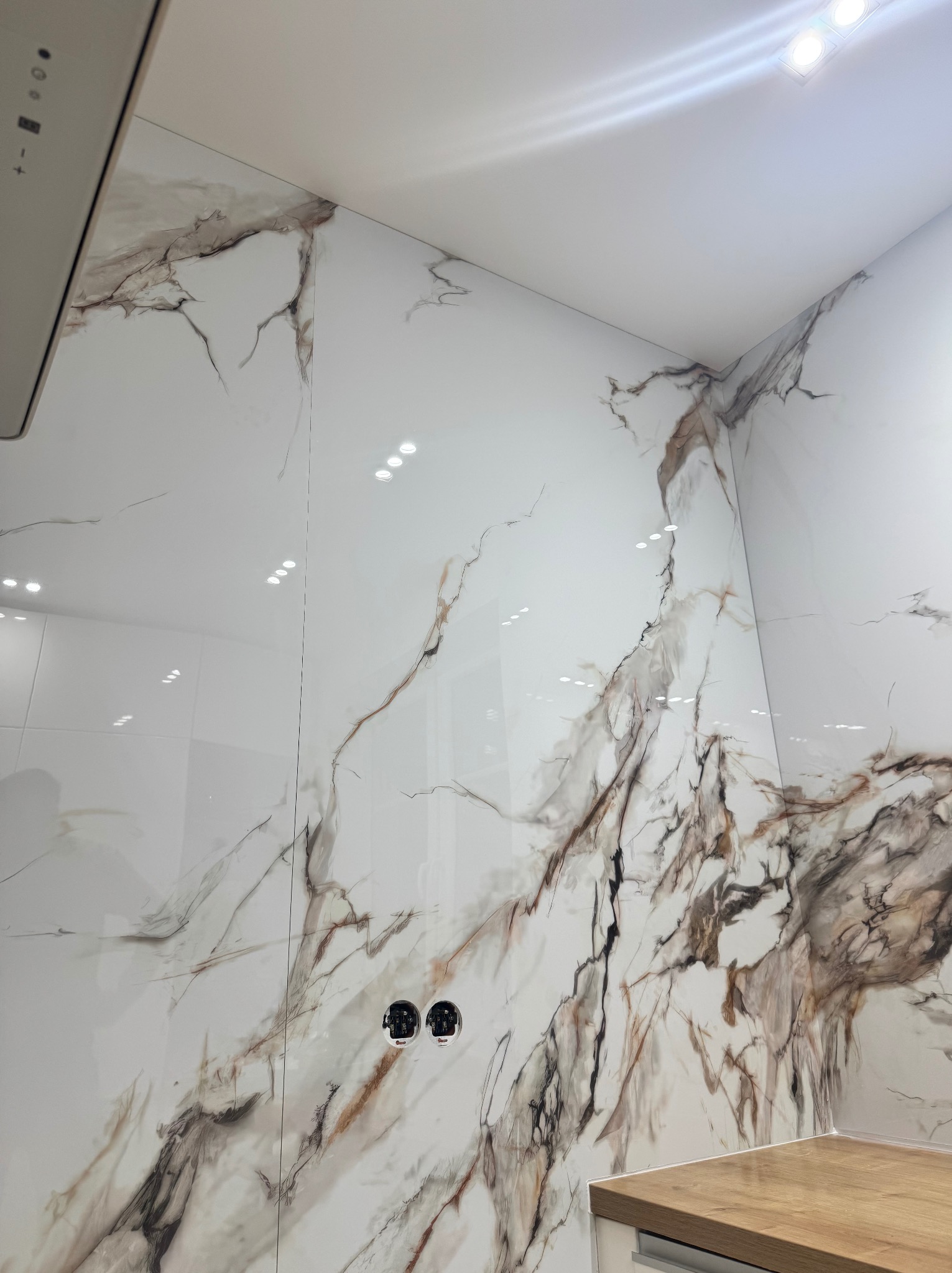 Białe płytki imitujące marmur z brązowymi żyłami na ścianie łazienki, widoczne gniazdka elektryczne i fragment blatu.