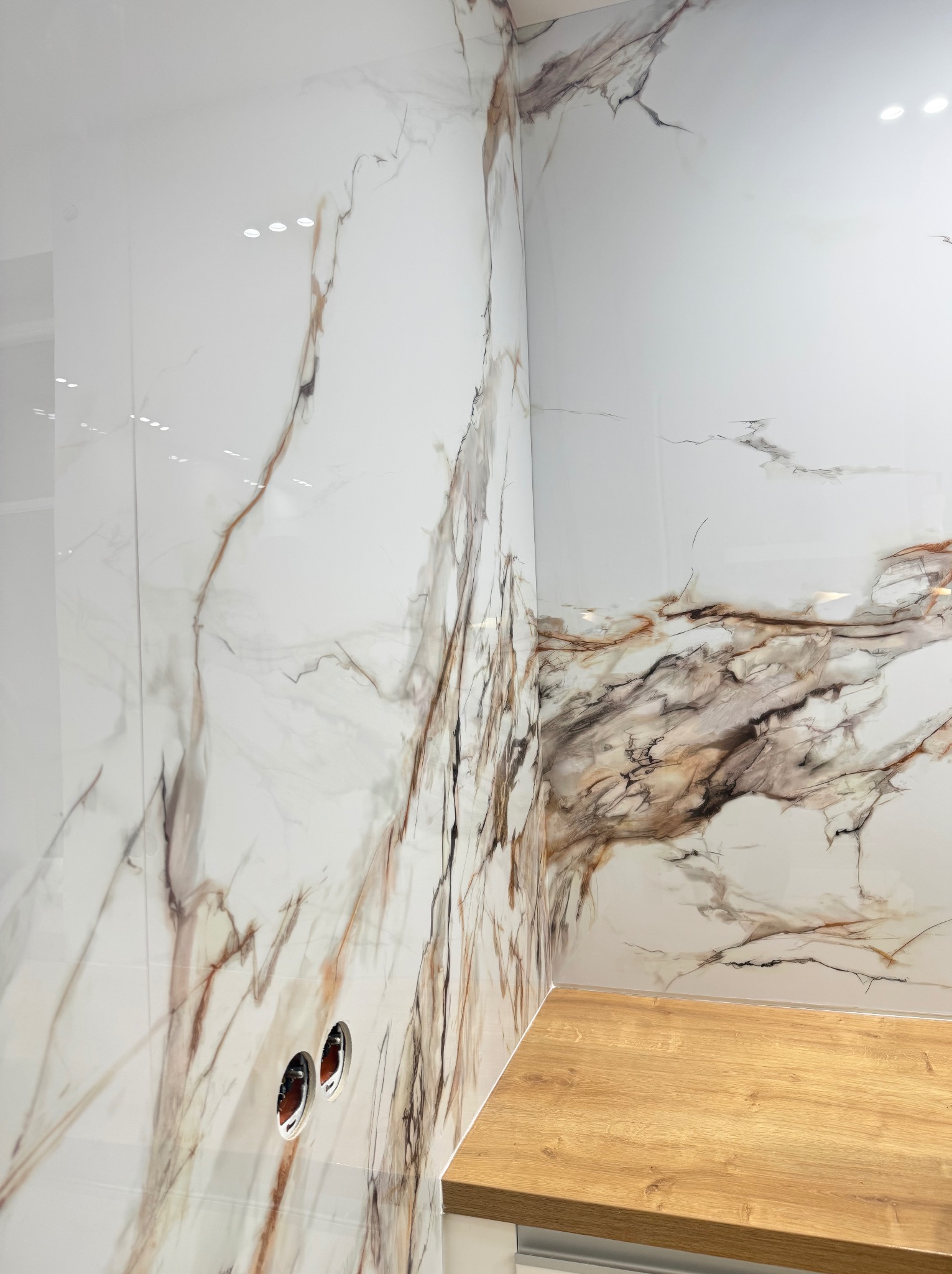 Białe, marmurowe płytki ścienne z brązowymi żyłami w rogu łazienki, widoczne dwa otwory na instalacje i fragment drewnianego blatu.