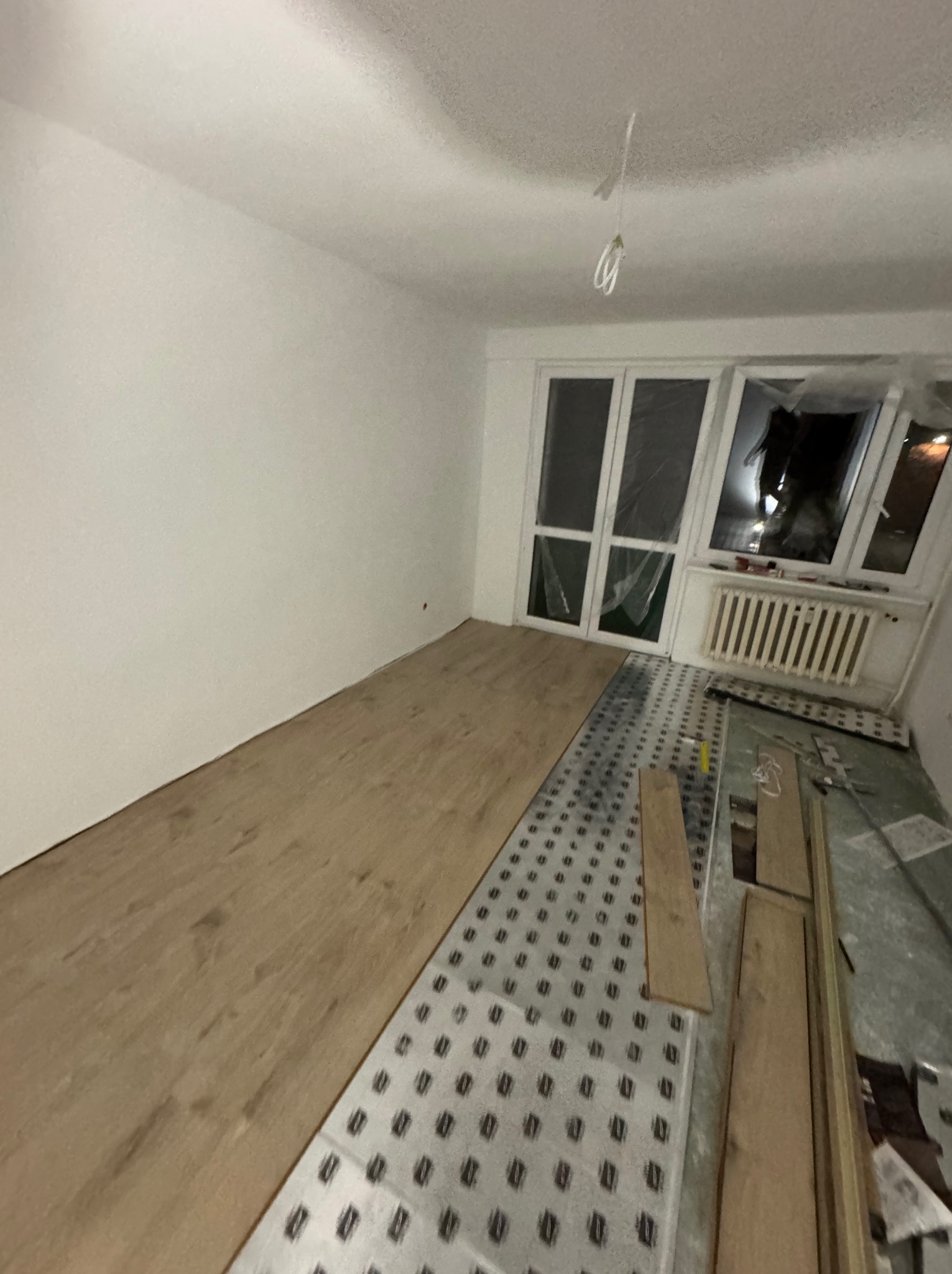 Fragment pokoju podczas układania paneli podłogowych; widoczna podkładka izolacyjna z wzorem, częściowo ułożone panele w kolorze jasnego drewna oraz okno balkonowe zabezpieczone folią malarską.