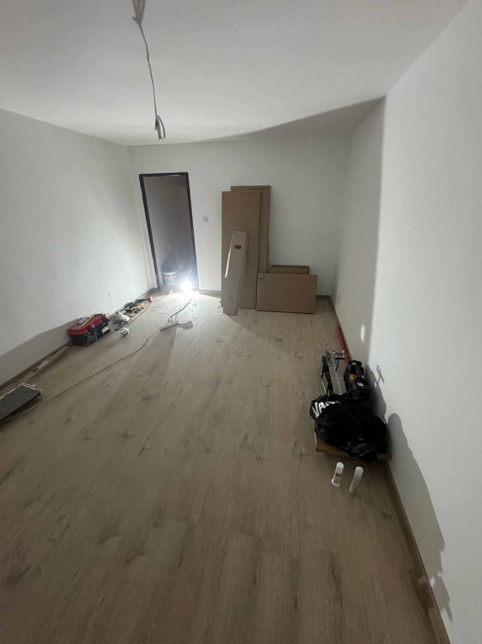 Pokój w trakcie remontu: świeżo położona podłoga z paneli, białe ściany, kartony i narzędzia budowlane, widoczne kable ze ściany.