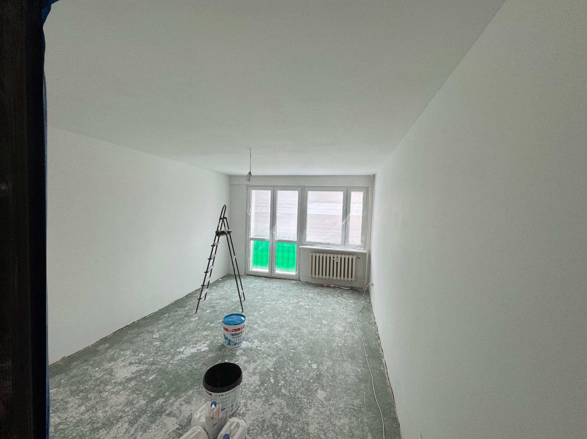 Pokój w trakcie odświeżania, świeżo pomalowane na biało ściany, zabezpieczone okno folią, drabina, wiaderka z farbą i pobrudzona farbą podłoga.