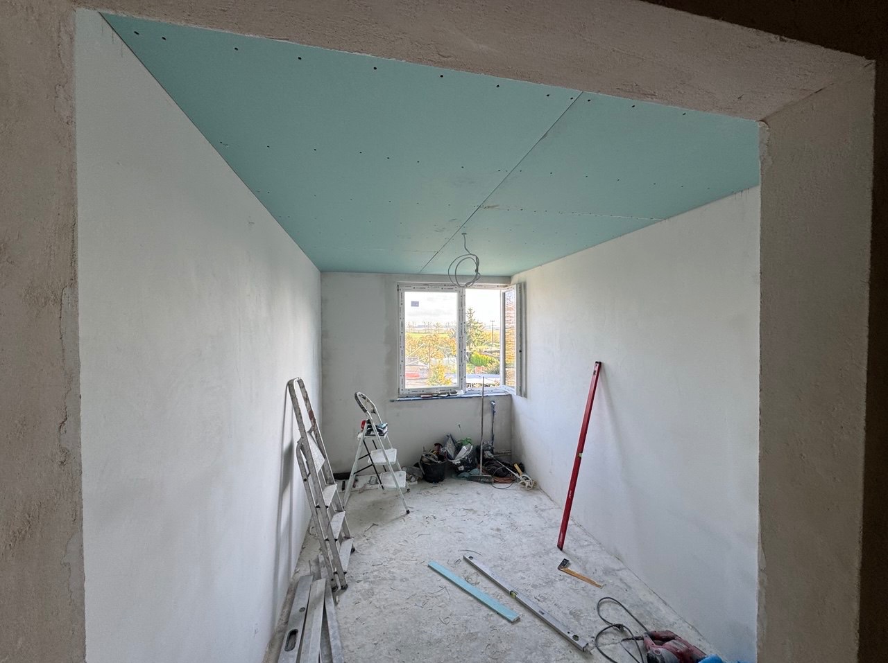 Wnętrze pokoju w trakcie remontu: ściany przygotowane do dalszych prac, sufit pokryty płytami kartonowo-gipsowymi w odcieniach błękitu, widoczne drabiny i narzędzia budowlane.