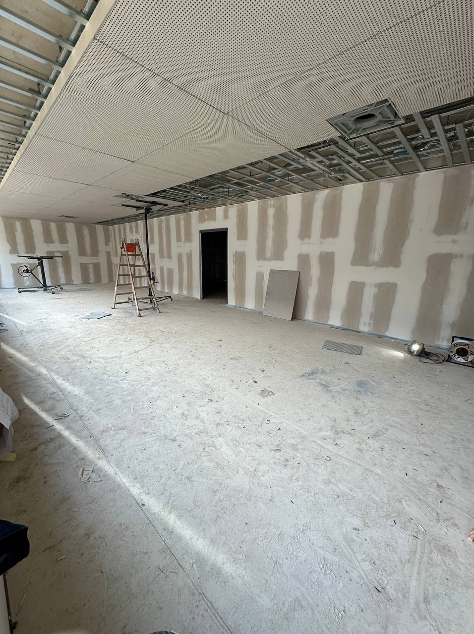 Wnętrze w Mieszkowicach w trakcie prac wykończeniowych: ściany z nałożonymi pasami szpachli, sufit podwieszany z perforowanych paneli, drabina i podnośnik do płyt na środku pomieszczenia.