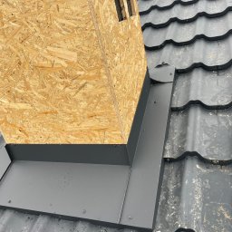PRO-DACH Blacharstwo Dekarstwo Adrian Bednarz - Szczegółowe ujęcie nowej, ciemnoszarej blachy okalającej komin z płyty OSB na dachu pokrytym dachówką ceramiczną. Widoczne krople wody na powierzchni dachu.