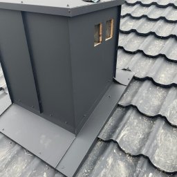 PRO-DACH Blacharstwo Dekarstwo Adrian Bednarz - Nowy, czarny komin z obróbką blacharską na dachu z metalowej dachówki, widoczny fragment roboczej rękawicy.