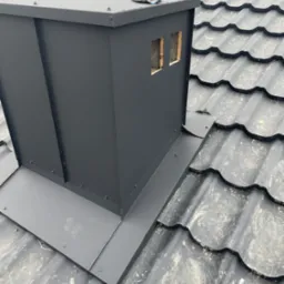 Nowy, czarny komin z obróbką blacharską na dachu z metalowej dachówki, widoczny fragment roboczej rękawicy.
