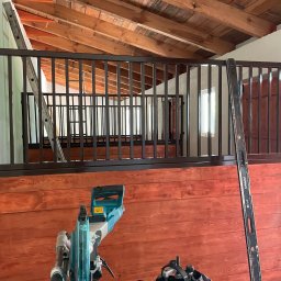 PRO-DACH Blacharstwo Dekarstwo Adrian Bednarz - Wnętrze poddasza z drewnianą konstrukcją dachu, widoczne barierki z metalowymi prętami, poziomica oparta o ścianę i pilarka ukośnica na pierwszym planie.