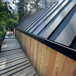 PRO-DACH Blacharstwo Dekarstwo Adrian Bednarz - Nowy, czarny, blaszany dach z wyraźnymi liniami łączeń, drewniana elewacja budynku, drewniany pomost roboczy i zielone drzewa w tle.