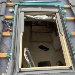 PRO-DACH Blacharstwo Dekarstwo Adrian Bednarz - Montaż okna dachowego na dachu pokrytym szarą dachówką, widoczne ocieplenie, narzędzia i drabina wewnątrz poddasza.