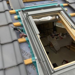 PRO-DACH Blacharstwo Dekarstwo Adrian Bednarz - Montaż okna dachowego w dachu z szarych dachówek, widoczna wkrętarka akumulatorowa i częściowo usunięte dachówki, wnętrze poddasza z widocznym stołem roboczym.