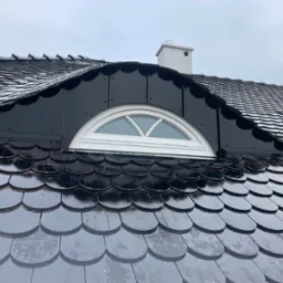 Dach pokryty błyszczącą, ciemną dachówką karpiówką, z lukarną w kształcie półkola i kominem w tle, lekko przyprószony śniegiem.