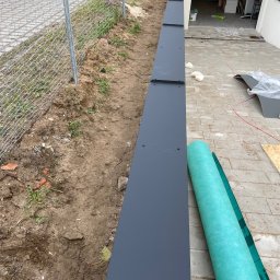 PRO-DACH Blacharstwo Dekarstwo Adrian Bednarz - Montaż obróbek blacharskich przy ogrodzeniu, widoczne elementy mocowane śrubami, zwój membrany dachowej, narzędzia i materiały budowlane w tle.