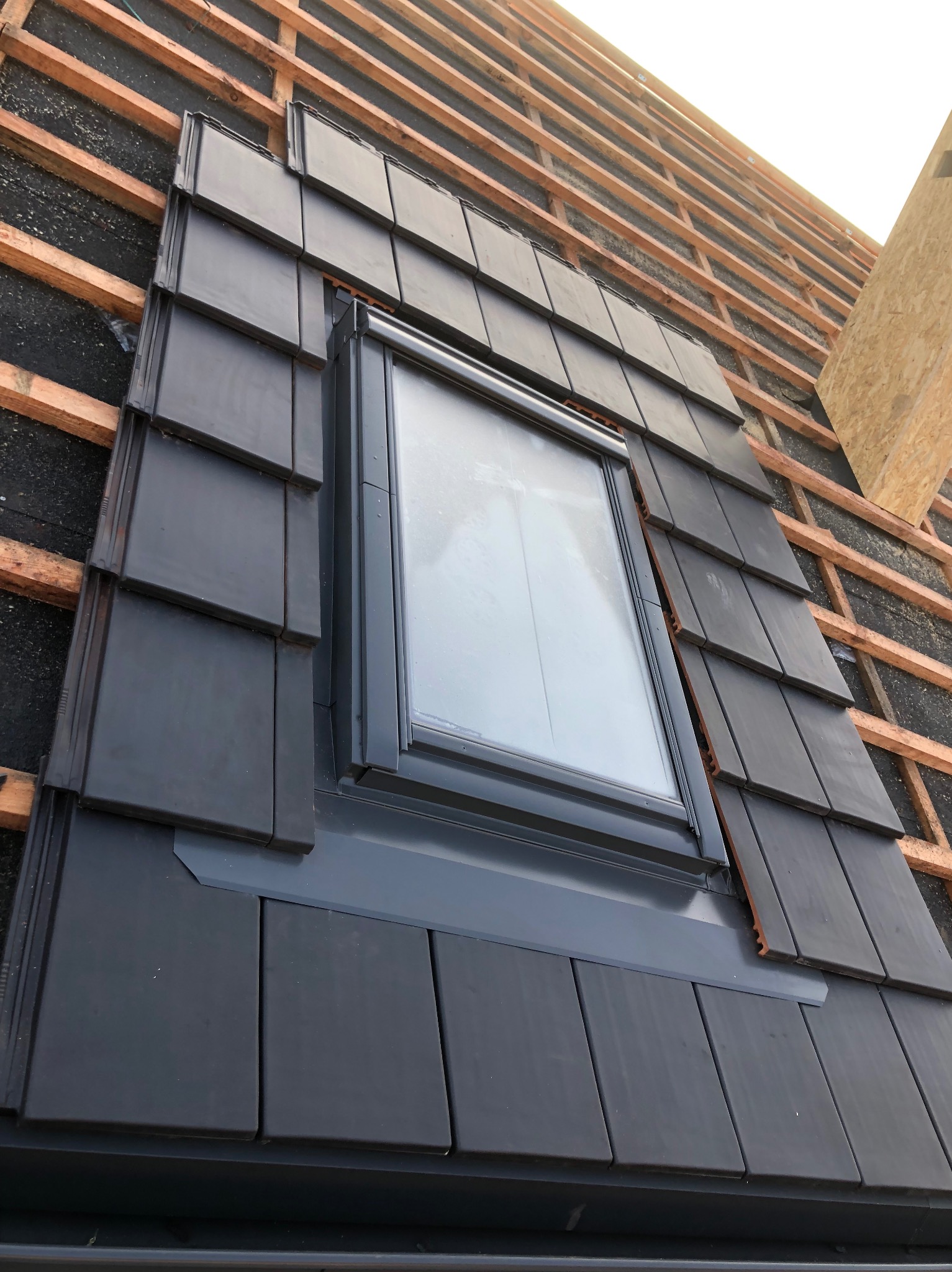 Montaż okna dachowego z szarą obróbką blacharską i czarną dachówką na konstrukcji drewnianej dachu.