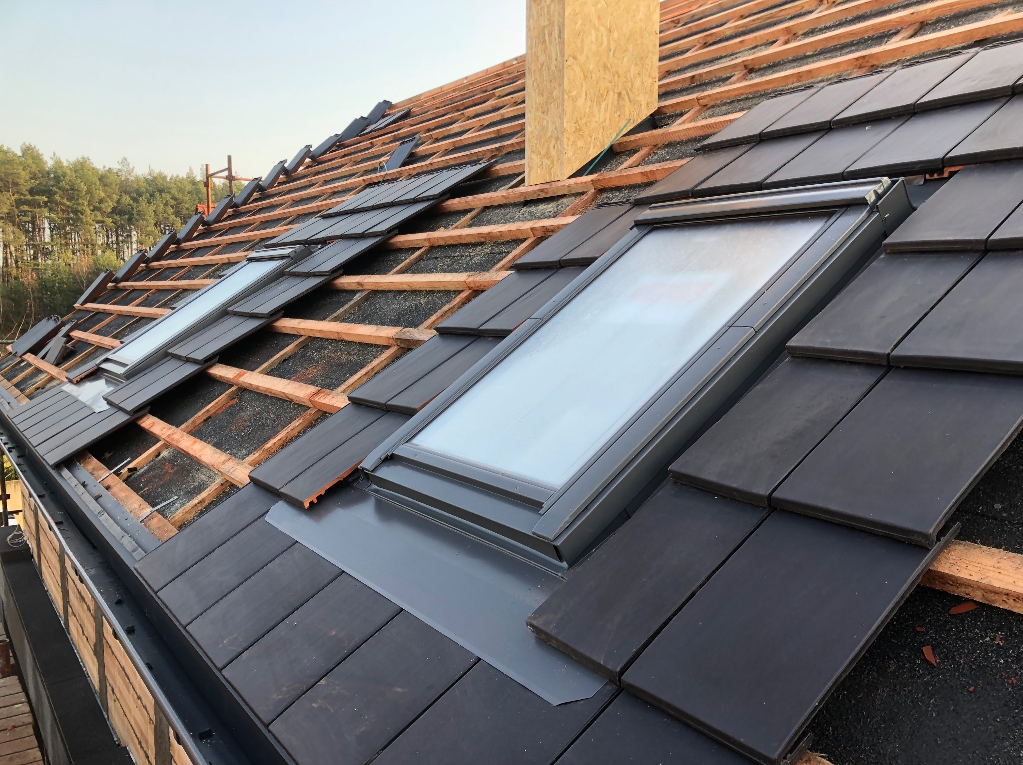 Montaż nowych dachówek wokół okien dachowych, widoczne drewniane łaty i fragment komina w surowym stanie.