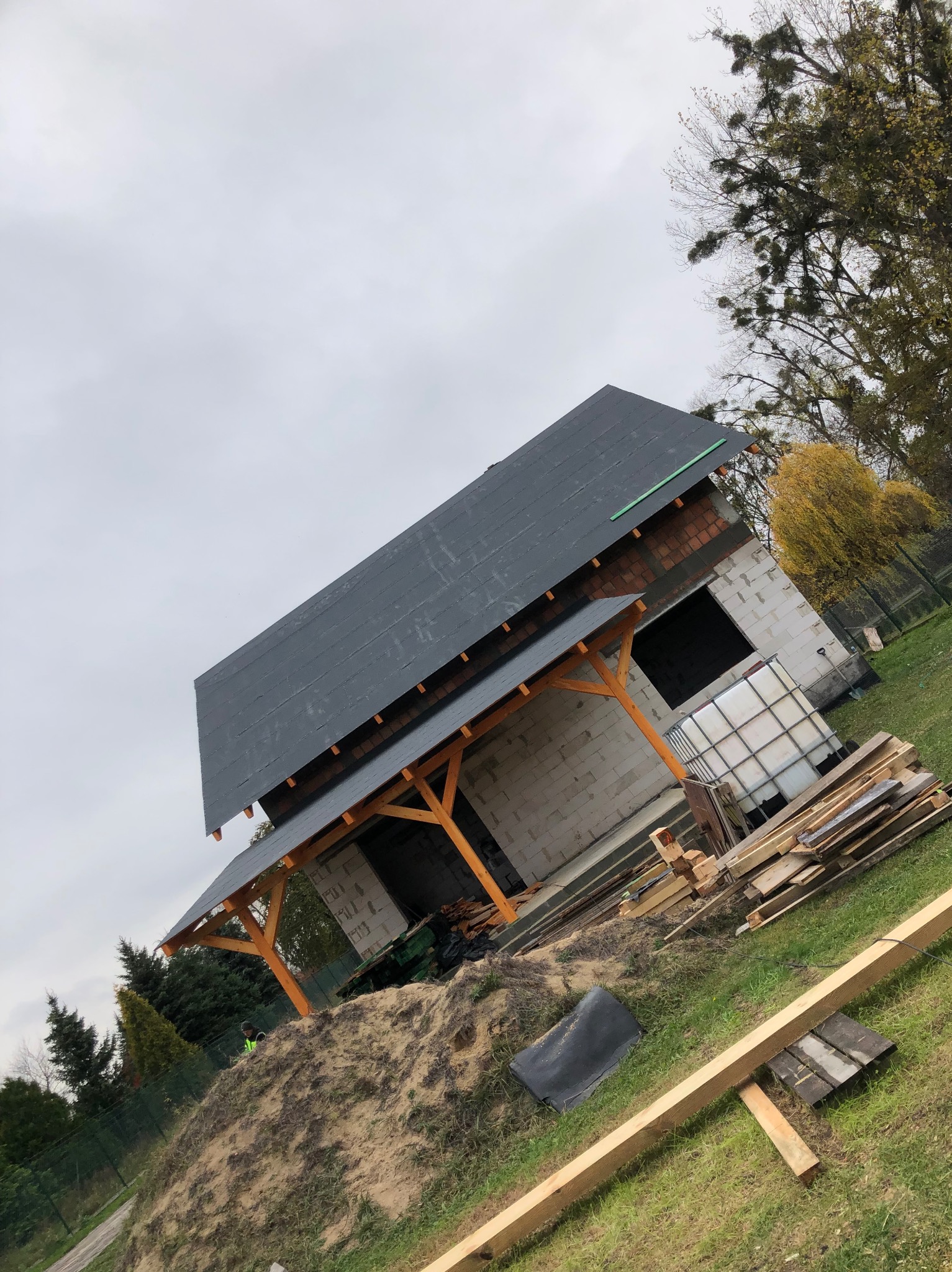 Budowa dachu z drewnianą konstrukcją i czarnym pokryciem, widoczne bloczki betonowe ścian i materiały budowlane na trawie, pochmurne niebo w tle.