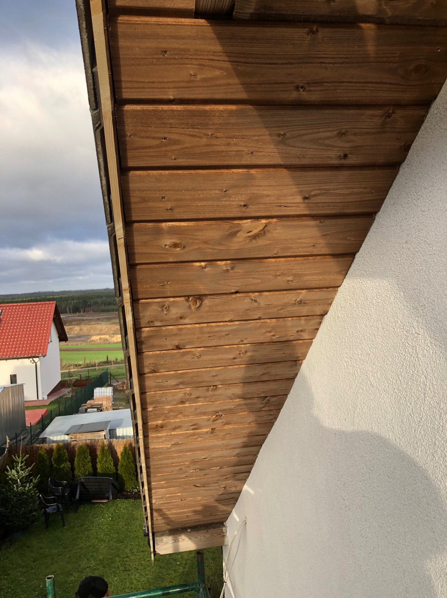 Drewniane deski dachowe z widocznymi śladami po sękach, świeżo zamontowane na poddaszu domu z białą elewacją i widokiem na ogród oraz okoliczne zabudowania.