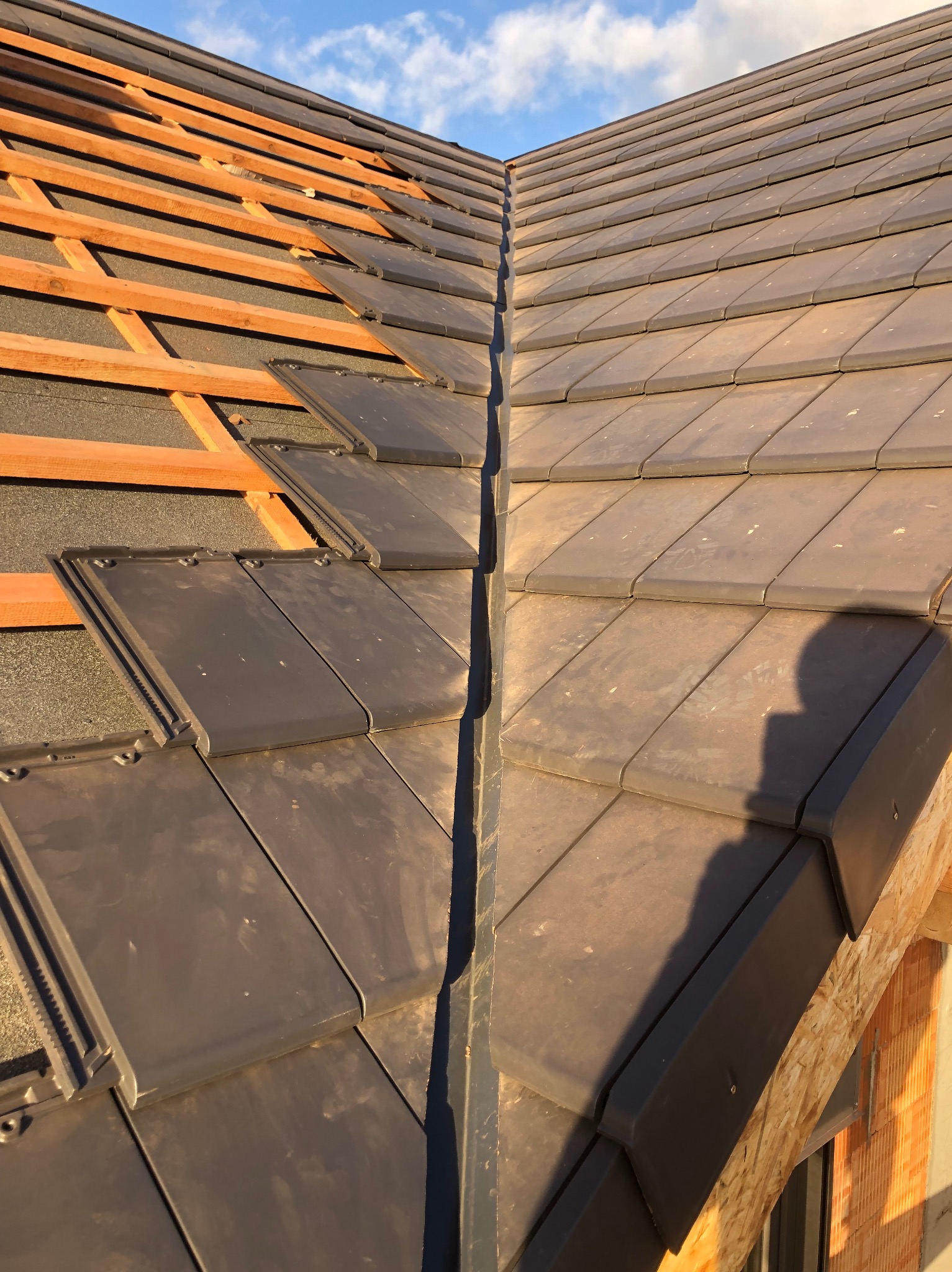 Fragment dachu w trakcie układania dachówki ceramicznej w kolorze grafitowym, widoczne łaty drewniane i membrana dachowa, w tle błękitne niebo z chmurami.