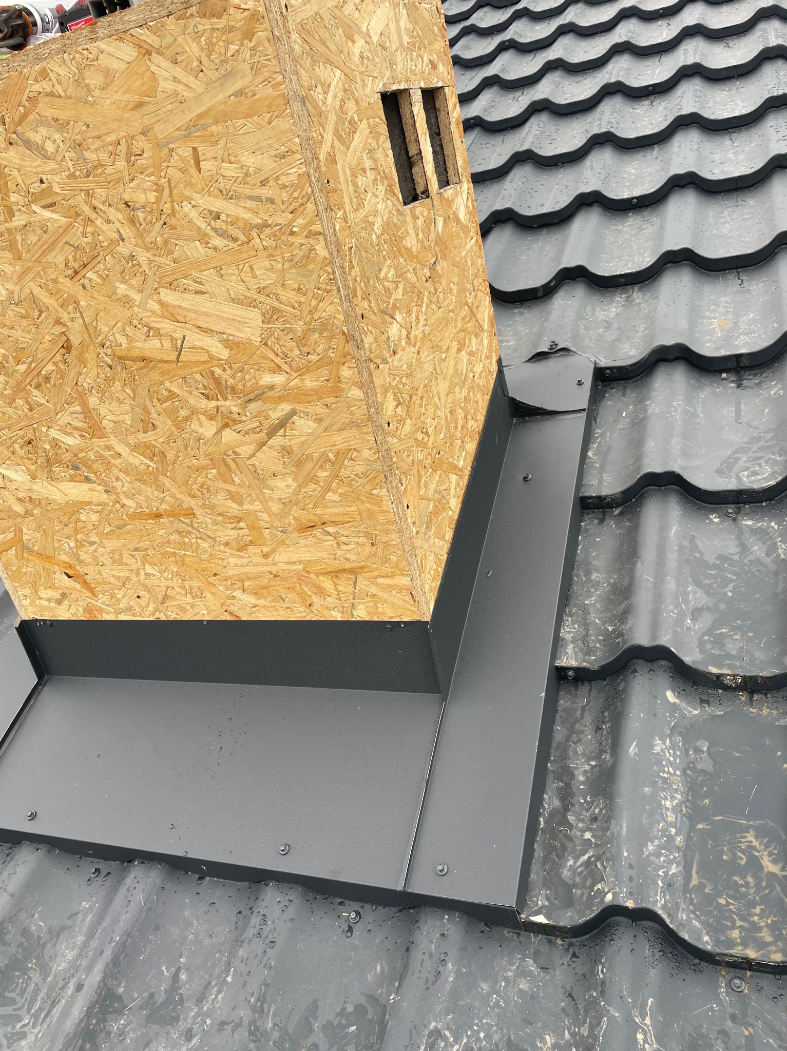 Szczegółowe ujęcie nowej, ciemnoszarej blachy okalającej komin z płyty OSB na dachu pokrytym dachówką ceramiczną. Widoczne krople wody na powierzchni dachu.