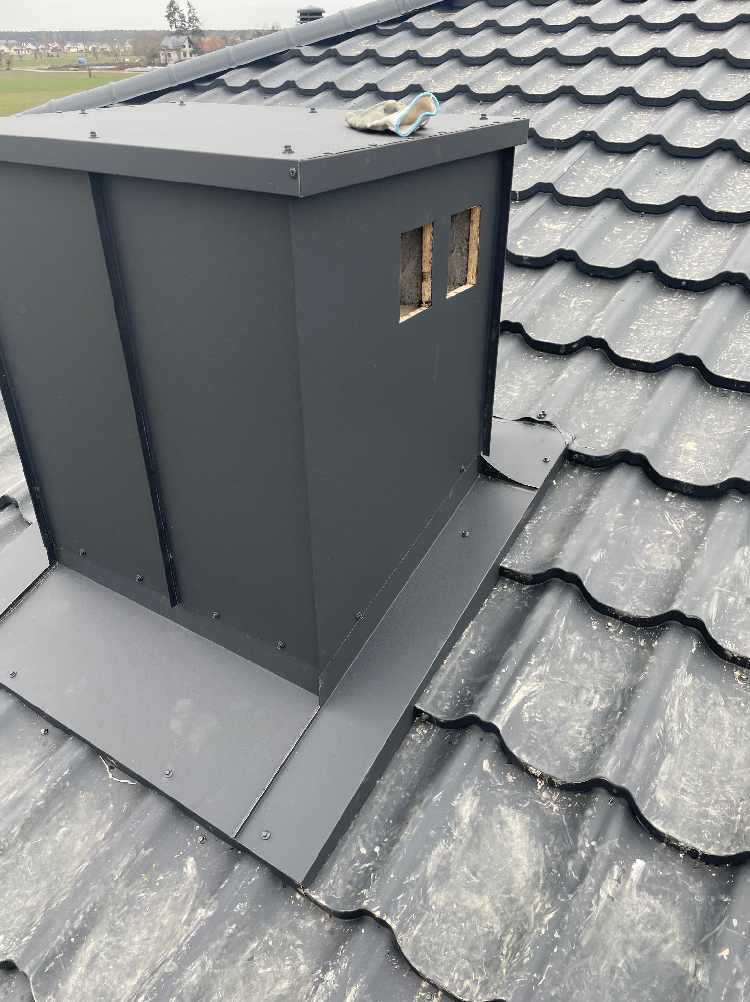 Nowy, czarny komin z obróbką blacharską na dachu z metalowej dachówki, widoczny fragment roboczej rękawicy.