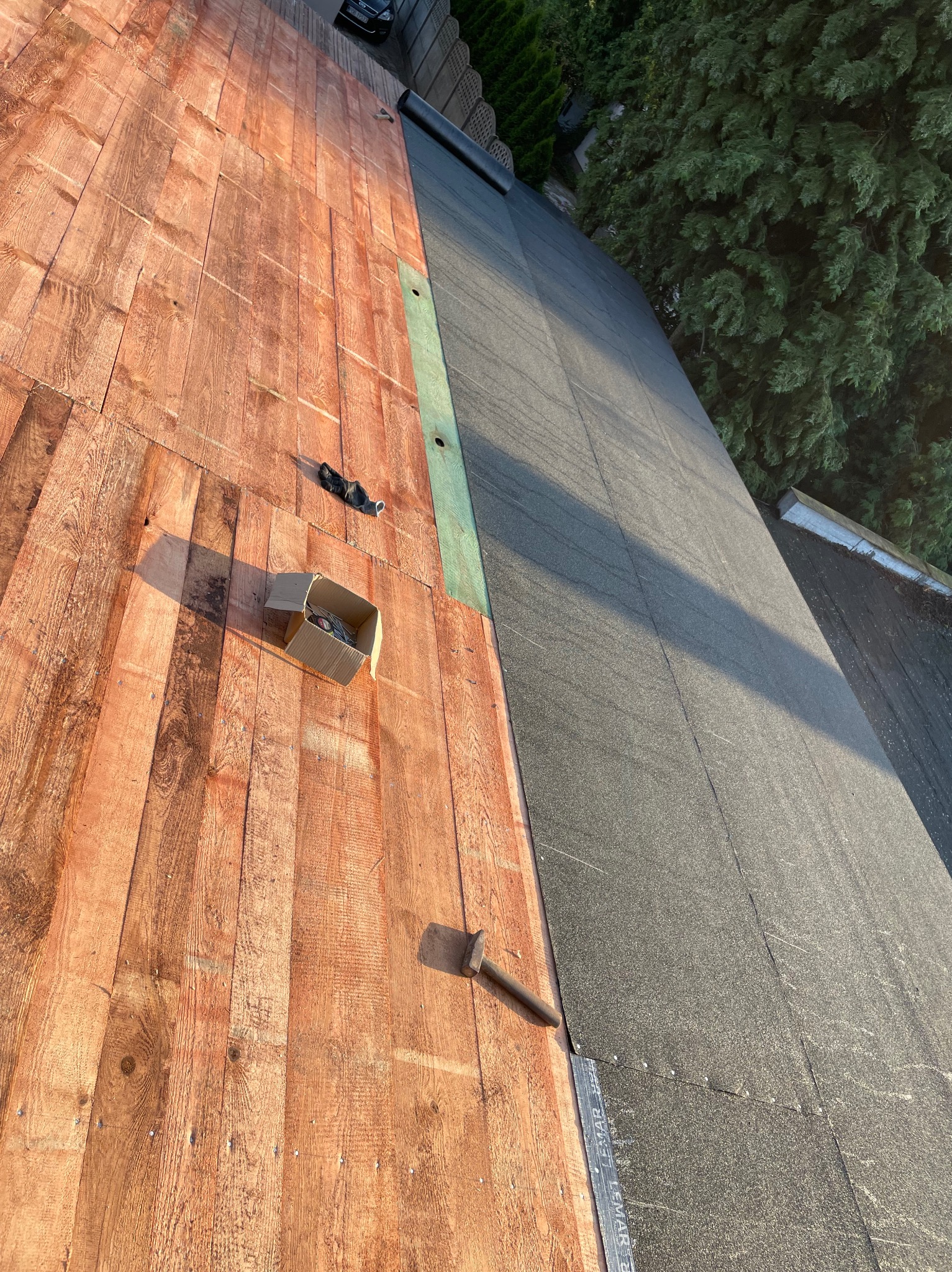 Połowa dachu pokryta deskami, druga połowa czarną papą, widoczny młotek, karton z gwoździami i rękawica robocza.