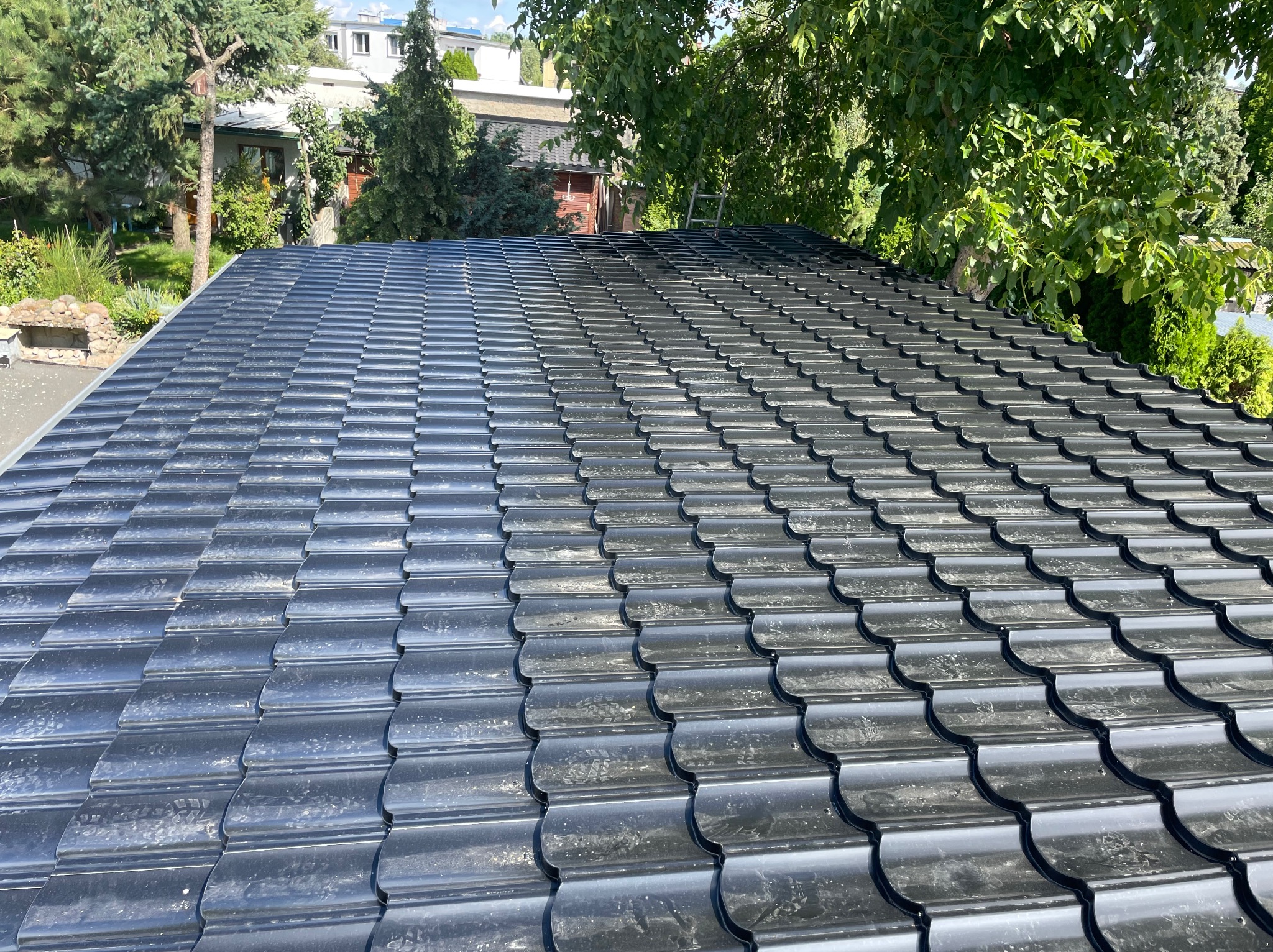 Nowy, błyszczący, grafitowy dach z dachówki ceramicznej, częściowo zabrudzony pyłem budowlanym, na tle zieleni drzew i zabudowań.