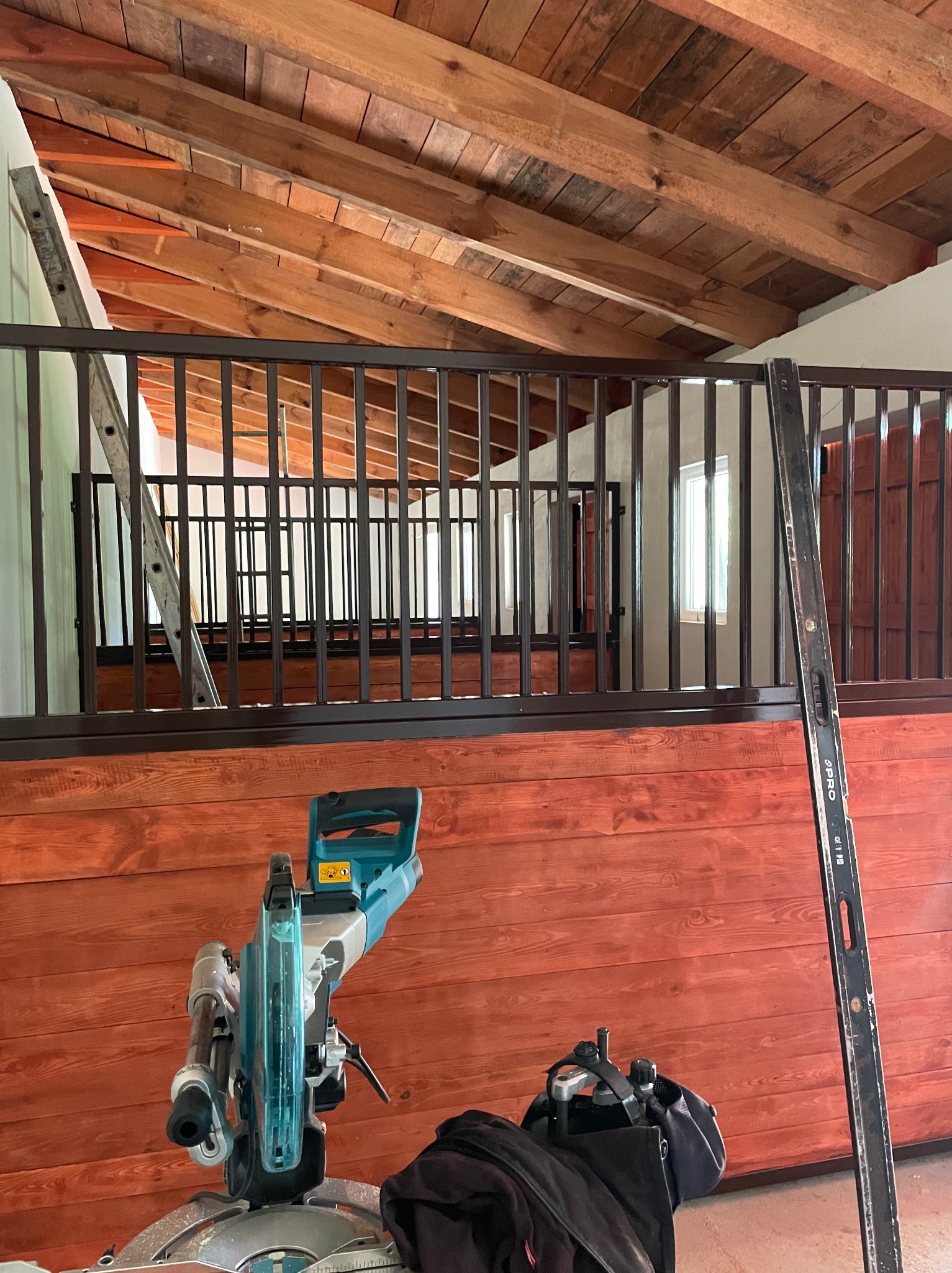 Wnętrze poddasza z drewnianą konstrukcją dachu, widoczne barierki z metalowymi prętami, poziomica oparta o ścianę i pilarka ukośnica na pierwszym planie.