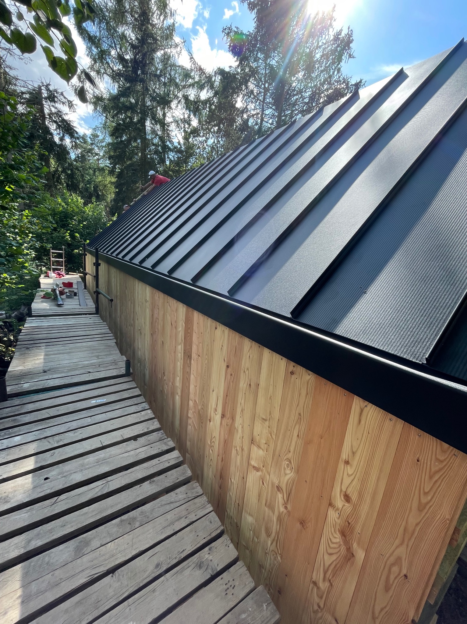 Nowy, czarny, blaszany dach z wyraźnymi liniami łączeń, drewniana elewacja budynku, drewniany pomost roboczy i zielone drzewa w tle.