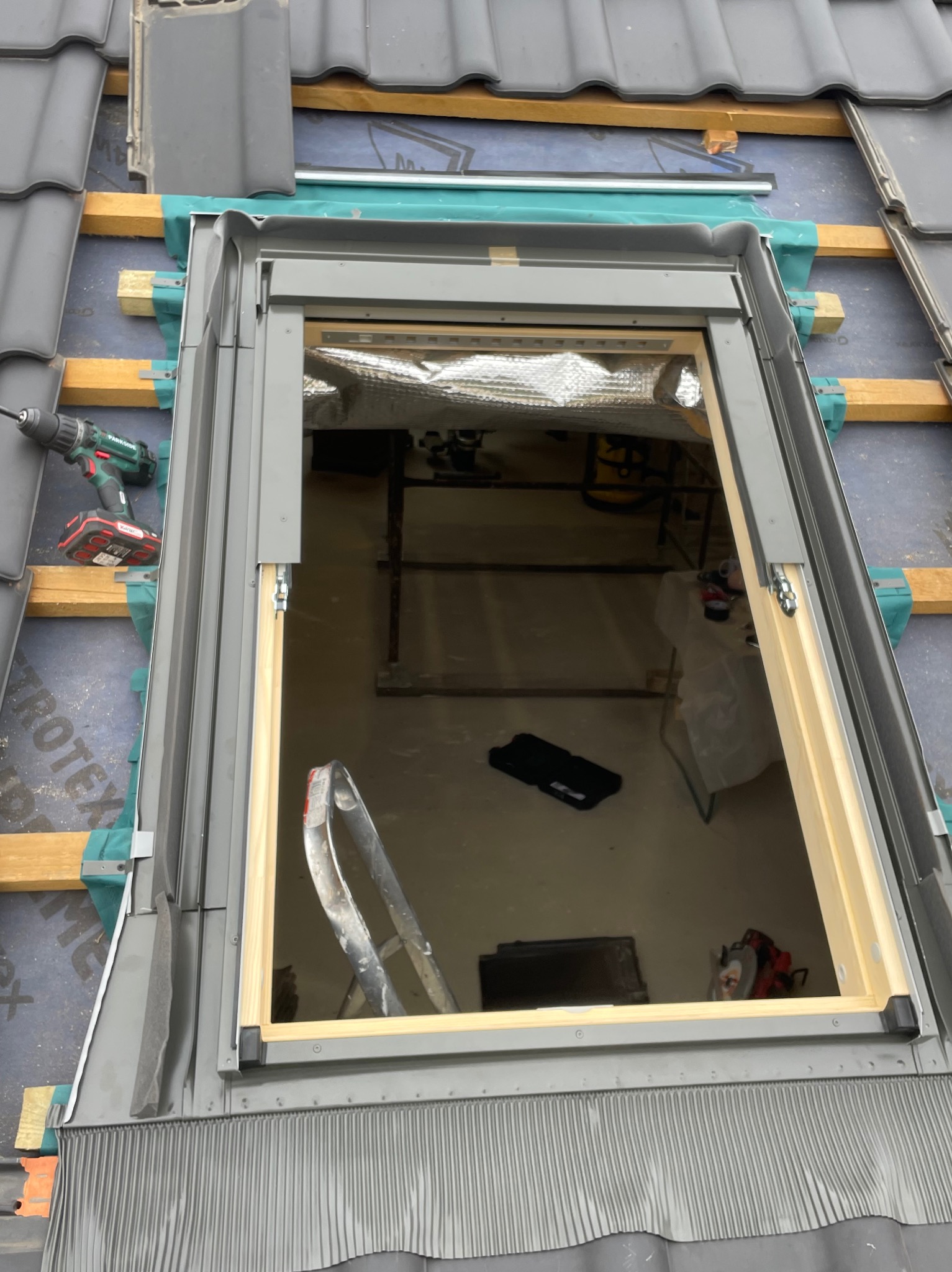 Montaż okna dachowego na dachu pokrytym szarą dachówką, widoczne ocieplenie, narzędzia i drabina wewnątrz poddasza.