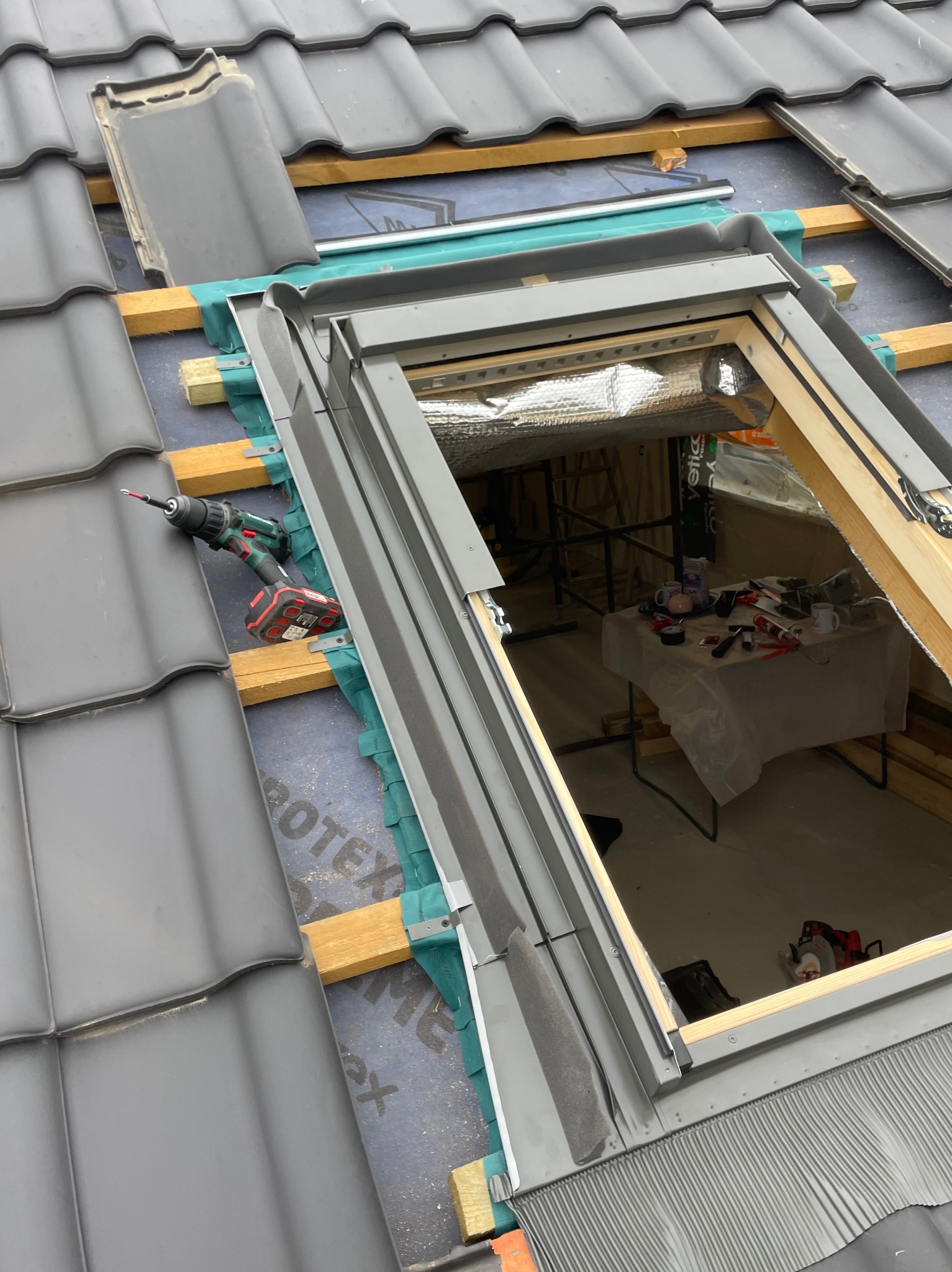 Montaż okna dachowego w dachu z szarych dachówek, widoczna wkrętarka akumulatorowa i częściowo usunięte dachówki, wnętrze poddasza z widocznym stołem roboczym.