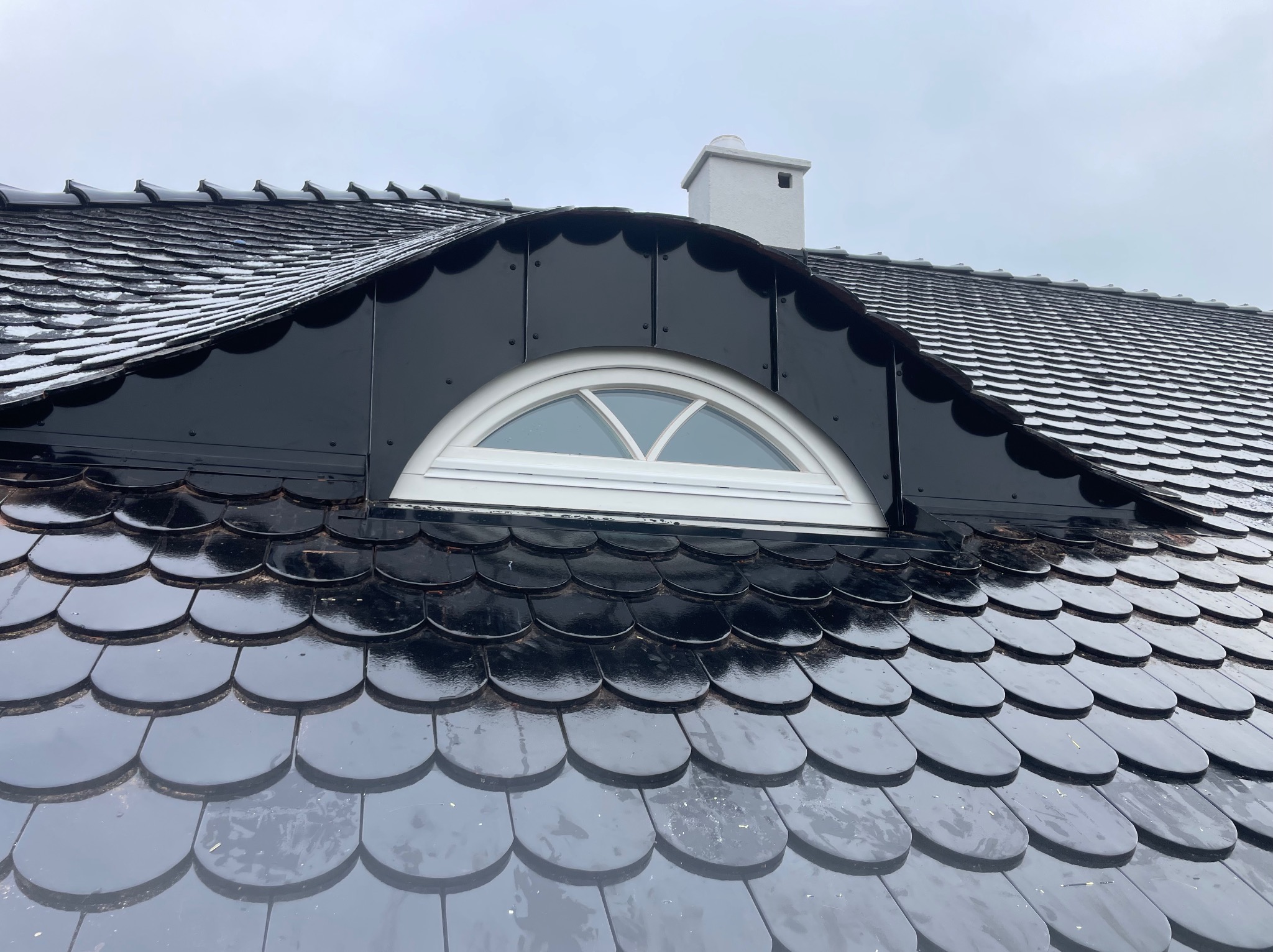 Dach pokryty błyszczącą, ciemną dachówką karpiówką, z lukarną w kształcie półkola i kominem w tle, lekko przyprószony śniegiem.