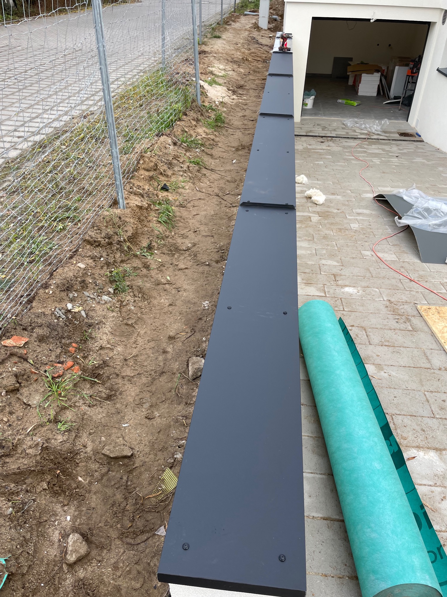 Montaż obróbek blacharskich przy ogrodzeniu, widoczne elementy mocowane śrubami, zwój membrany dachowej, narzędzia i materiały budowlane w tle.